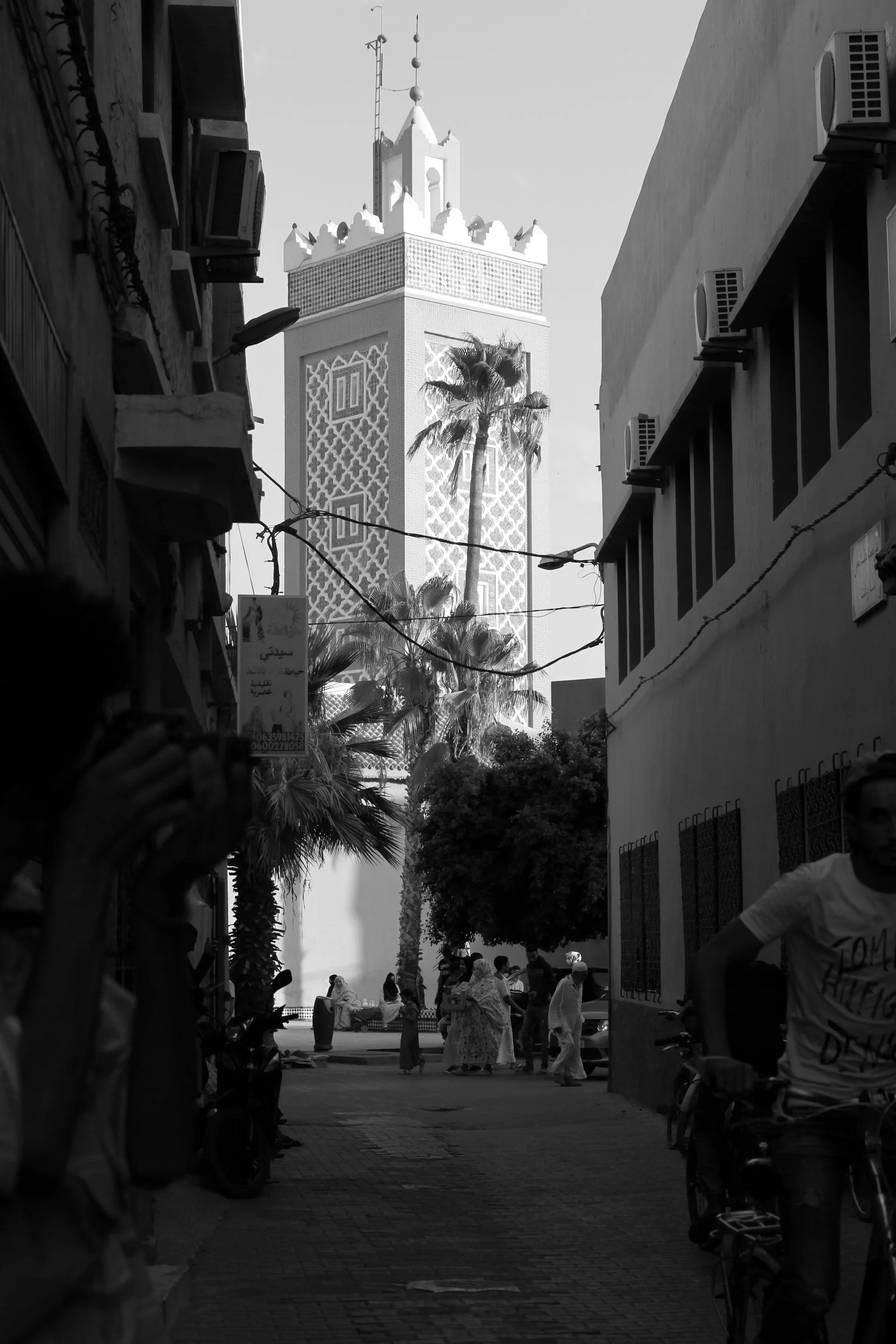 A crowded street scene with people walking and riding bicycles, tall palm trees, and a large ornate tower in the background, depicted in black and white.
