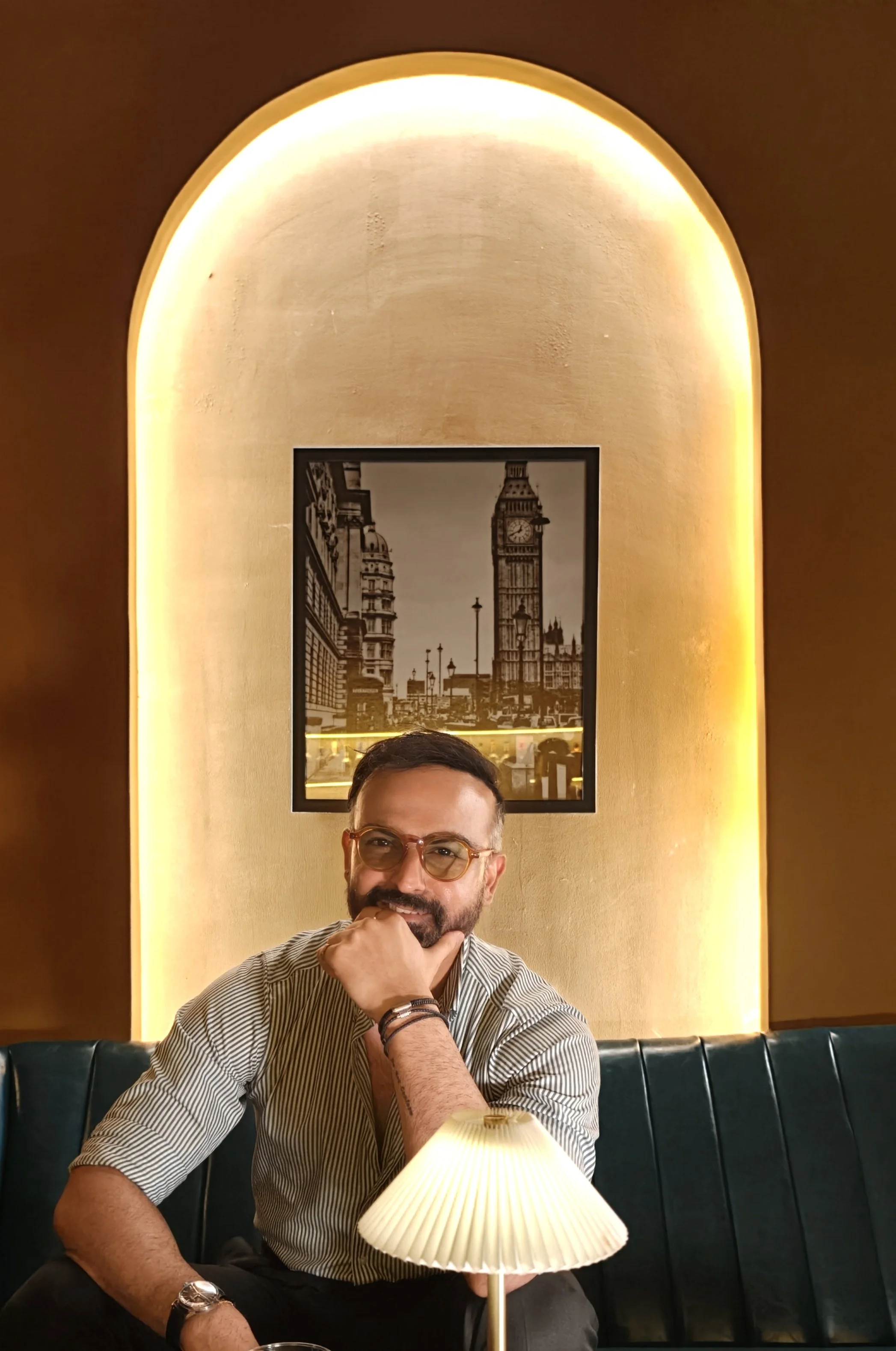 A man with glasses and a beard sitting at a table in a restaurant, with his hand on his chin, in front of a lamp, with a black and white picture of Big Ben in the background