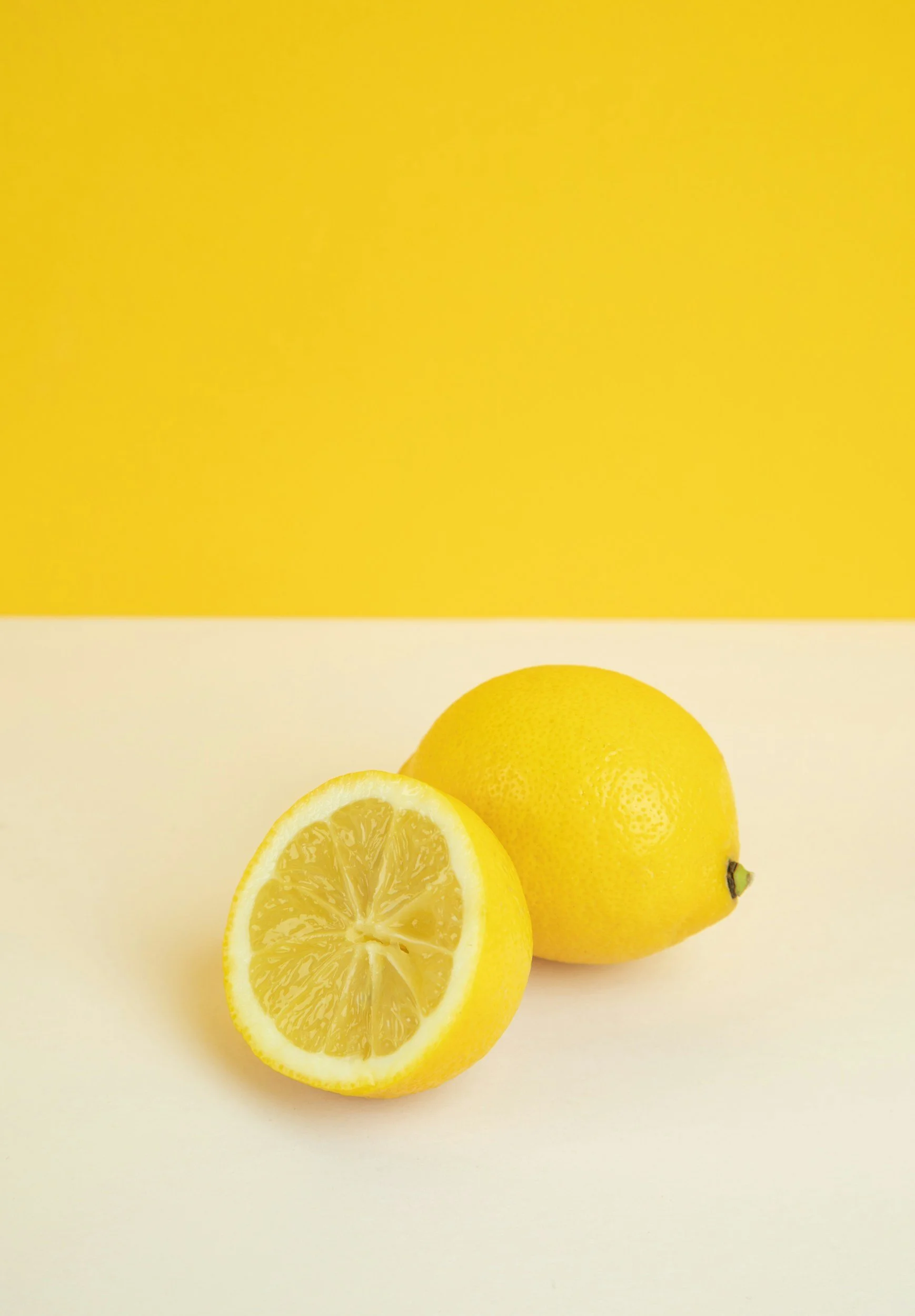 A whole yellow lemon and a lemon half with visible pulp on a light yellow surface and a yellow background.