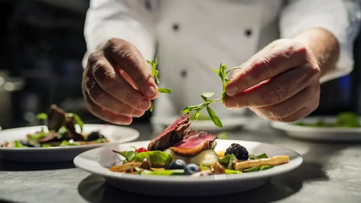 Chef garnishing a plated beef dish with herbs in a professional kitchen.
