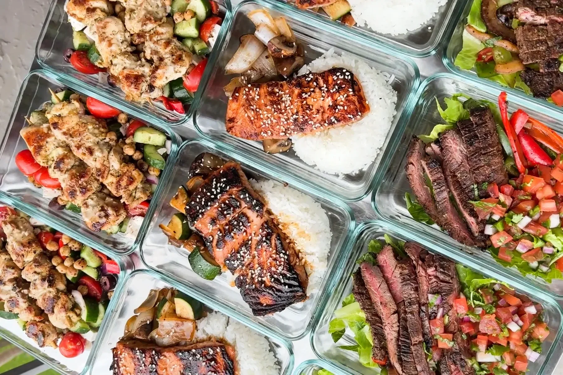 Assorted dishes in glass containers, including grilled salmon with sesame seeds over rice, grilled steak slices on a bed of vegetables, and other grilled meats and vegetable salads.