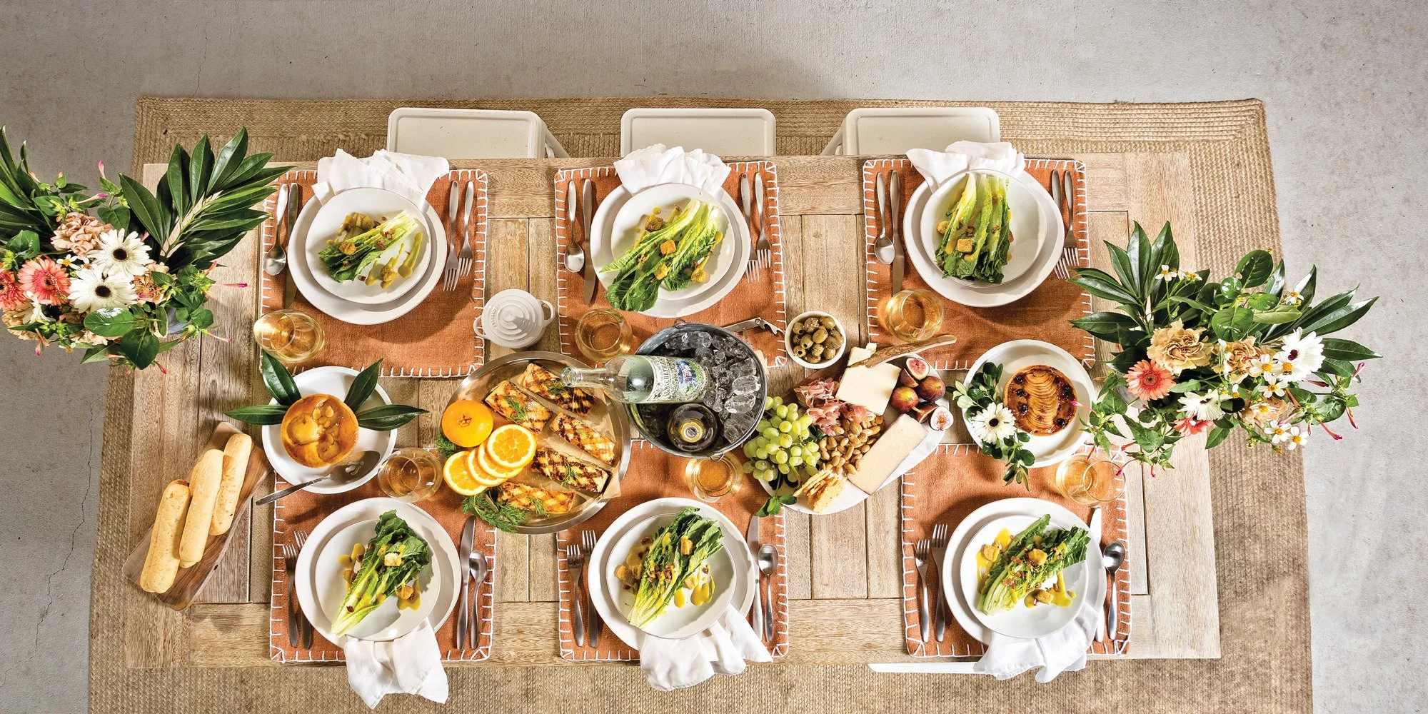 Overhead view of a decorated dining table set for a meal with plates of salads, breadsticks, cheese, figs, oranges, grapes, wine, and small bowls of olives and honey, with floral centerpieces on either side.