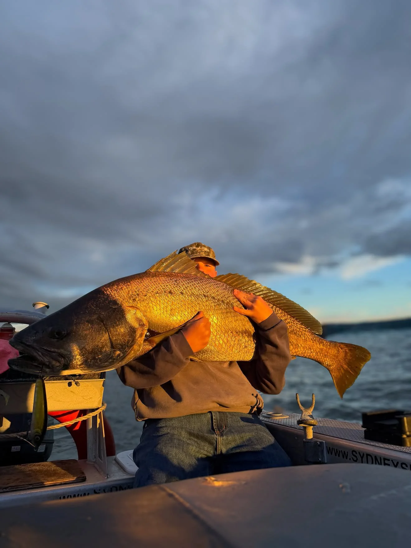 Person on boat holding a large fish against a cloudy sky, water in the background.