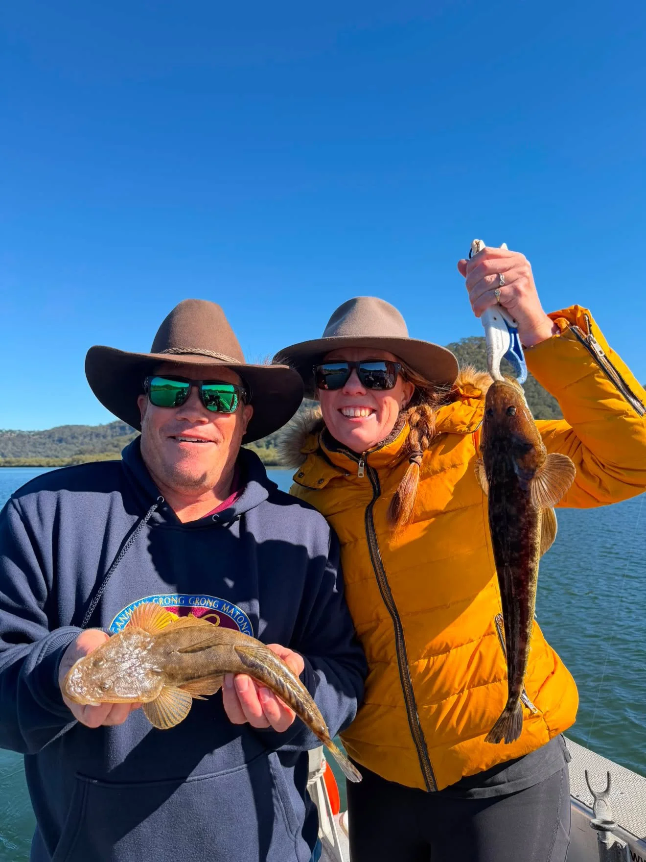 Two people wearing sunglasses and hats holding up fish they caught on a boat with a body of water and trees in the background.