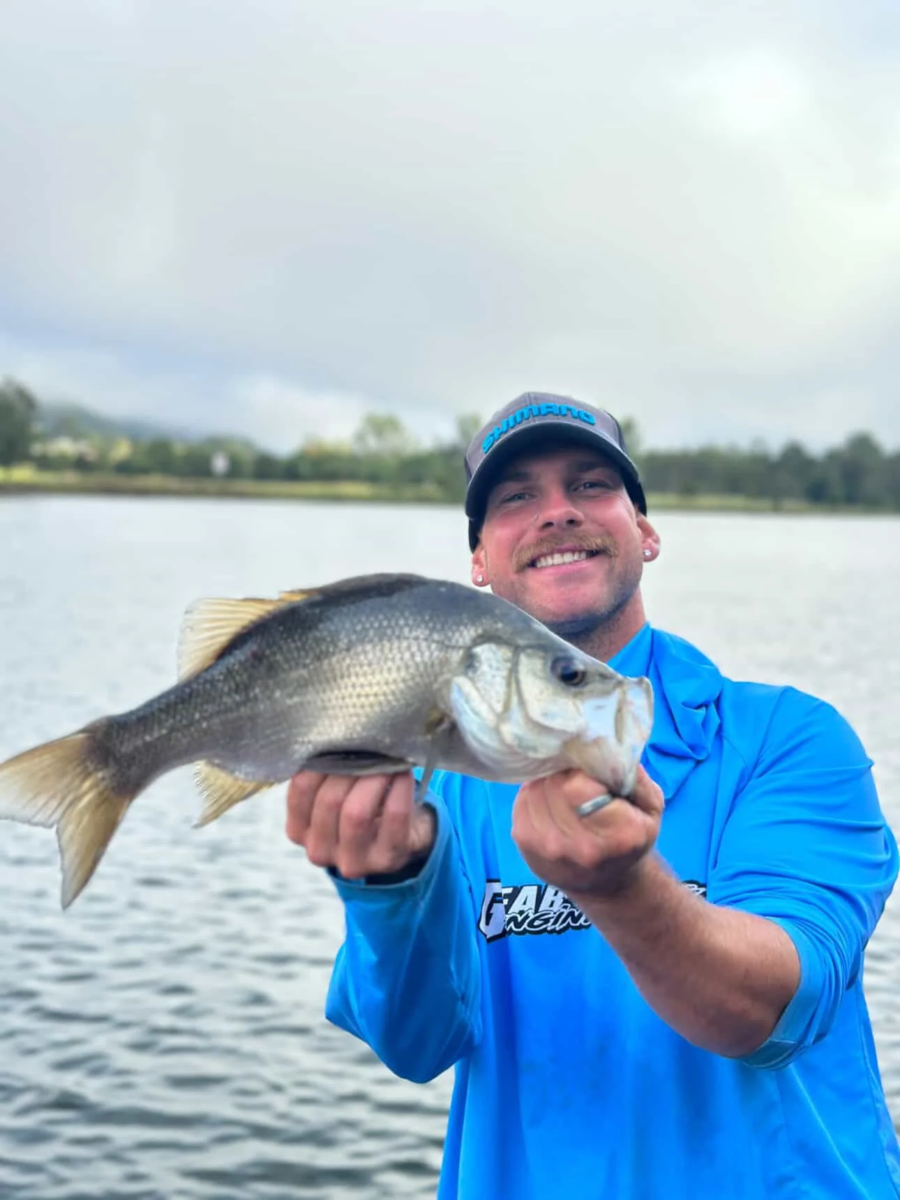 A man holding a large fish he caught while fishing on a lake, with a cloudy sky and trees in the background.