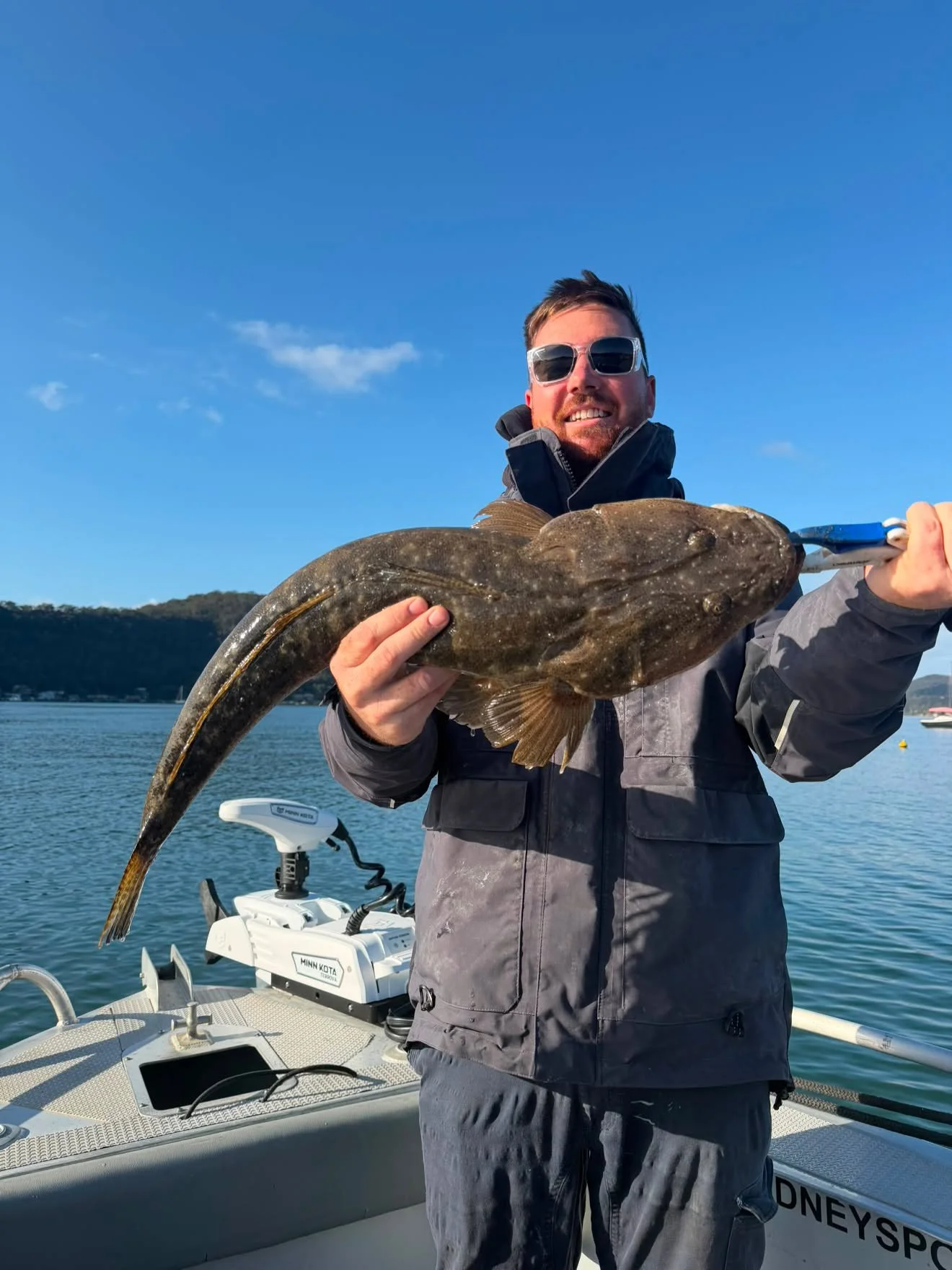 A smiling man wearing sunglasses and a gray jacket holding a large, flat fish on a boat. Water and hills are visible in the background.