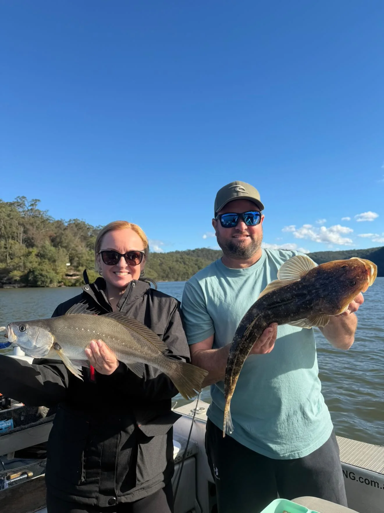 Two people holding fish on a boat with a body of water and hills in the background under clear blue sky. The woman on the left is wearing sunglasses and a black jacket, holding a large fish. The man on the right is wearing sunglasses, a cap, and a light blue shirt, smiling and holding a large dark fish.