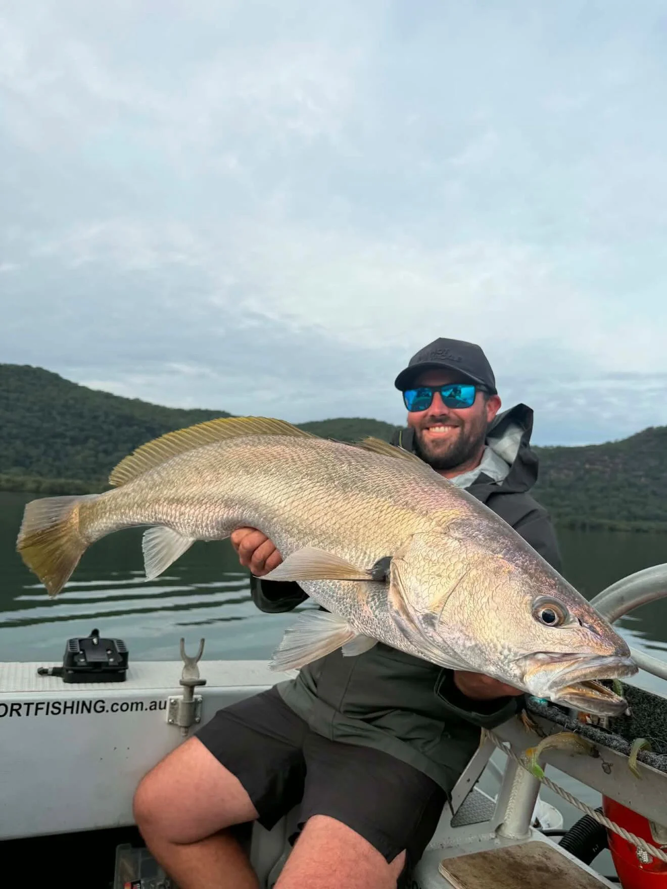 A man wearing sunglasses, a hat, and outdoor clothing sitting on a boat, holding a large fish and smiling. There are green hills and a cloudy sky in the background.