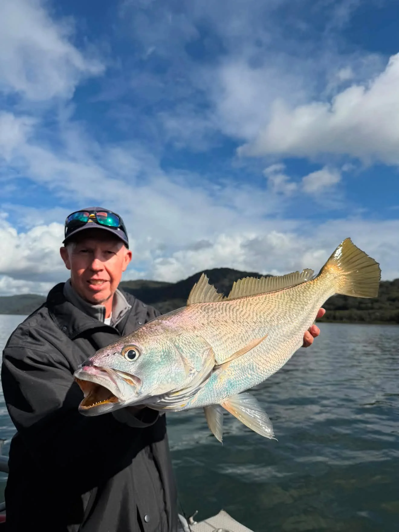 A man wearing a black jacket, sunglasses, and a cap is holding a large fish with a light-colored body and a big eye, on a background of water and cloudy sky.