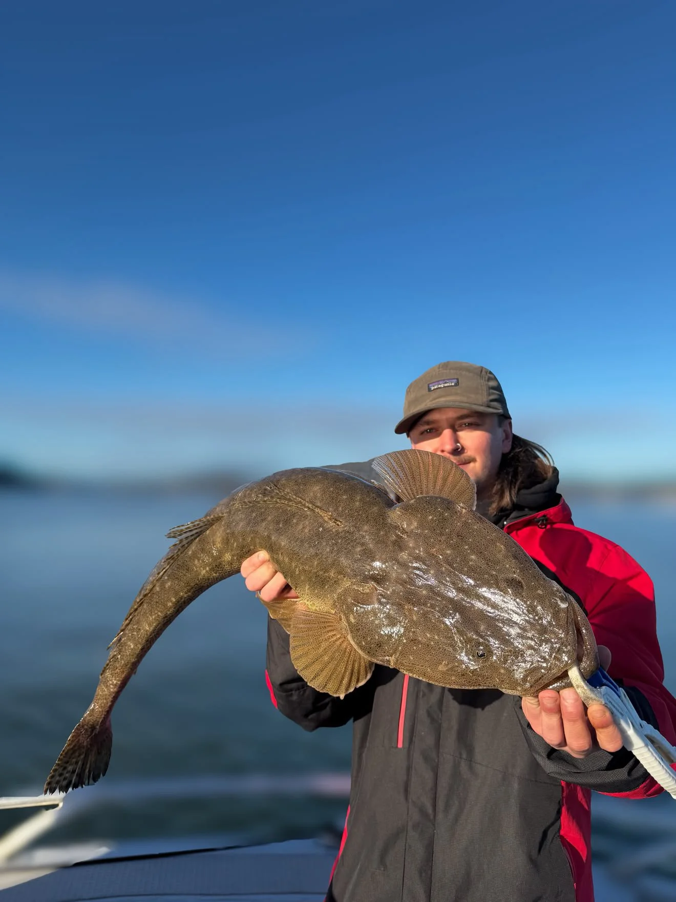 Person wearing a cap and red jacket holding a large fish on a boat with a blue sky in the background.