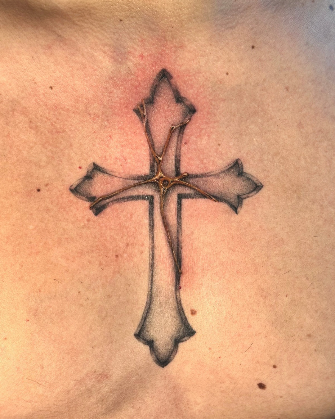 Tattoo of a cross with a floral or leaf-like design inside, outlined in black with shading, on skin.