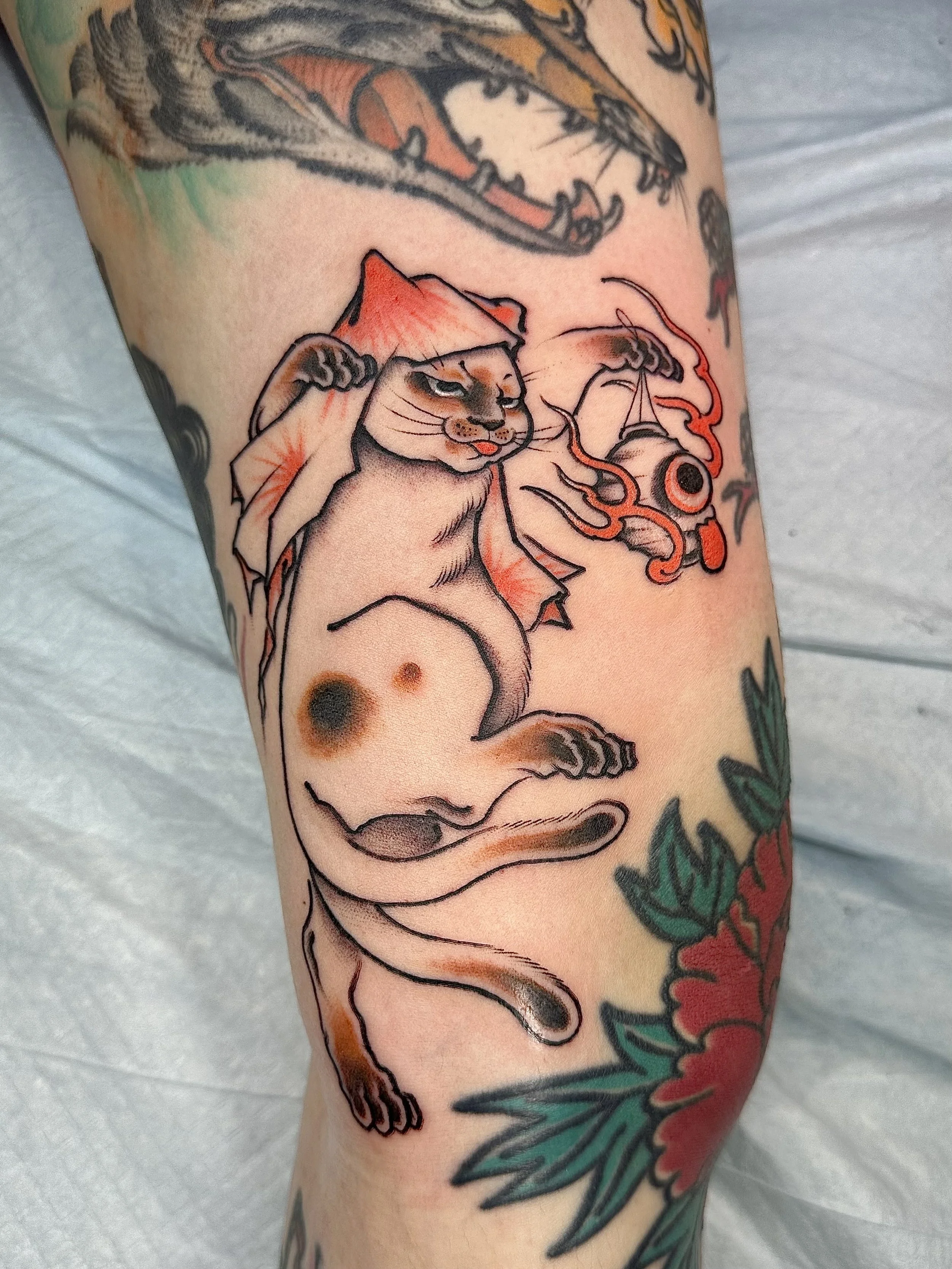 Colorful tattoo of a black cat with yellow eyes, holding a guitar, surrounded by orange flowers, with additional decorative elements like flames and a small cat face below the guitar.