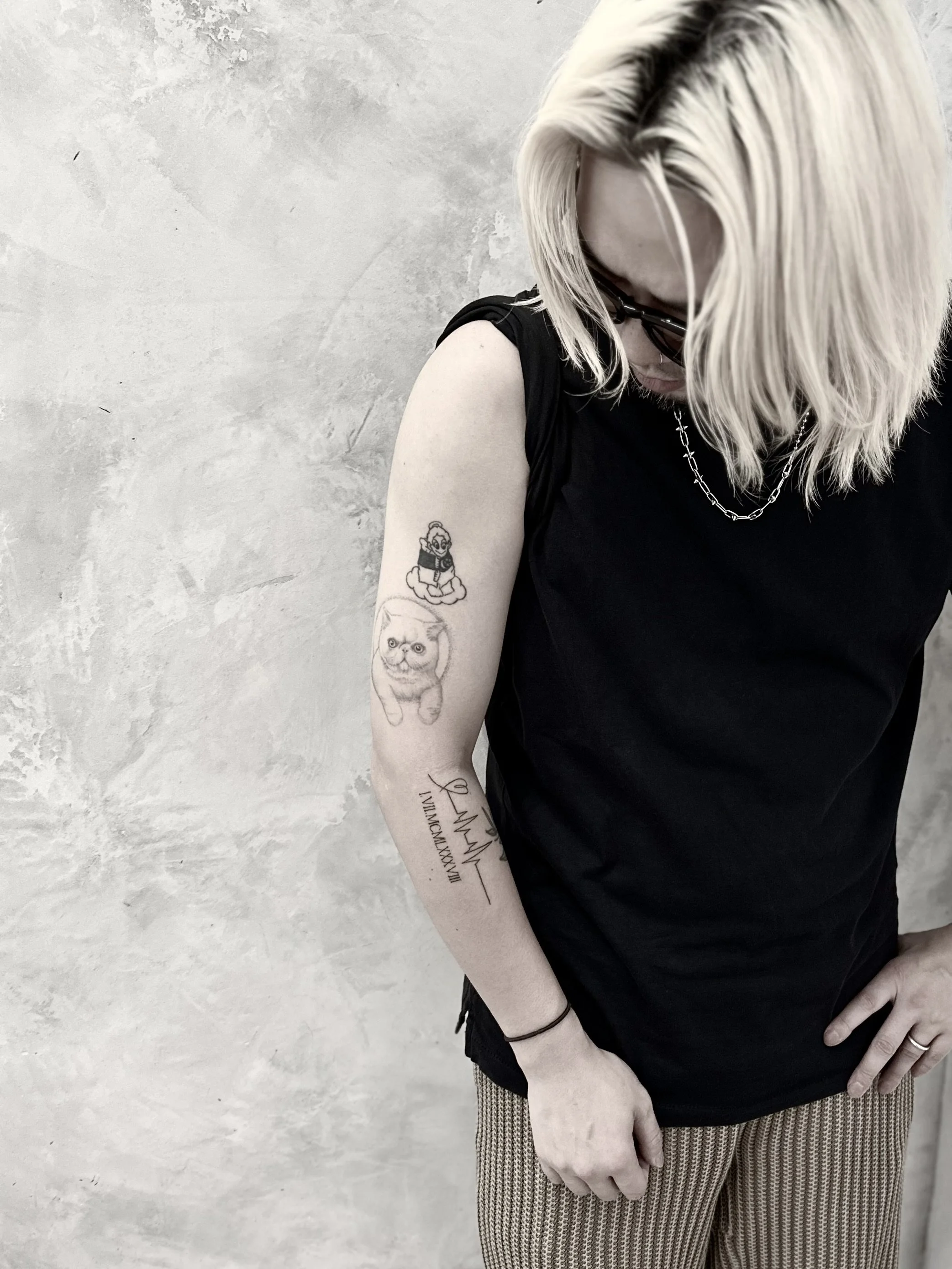 Person with blonde hair wearing glasses, a black sleeveless shirt, a silver chain necklace, and patterned pants, showcasing tattoos on the left arm, against a textured light gray background.