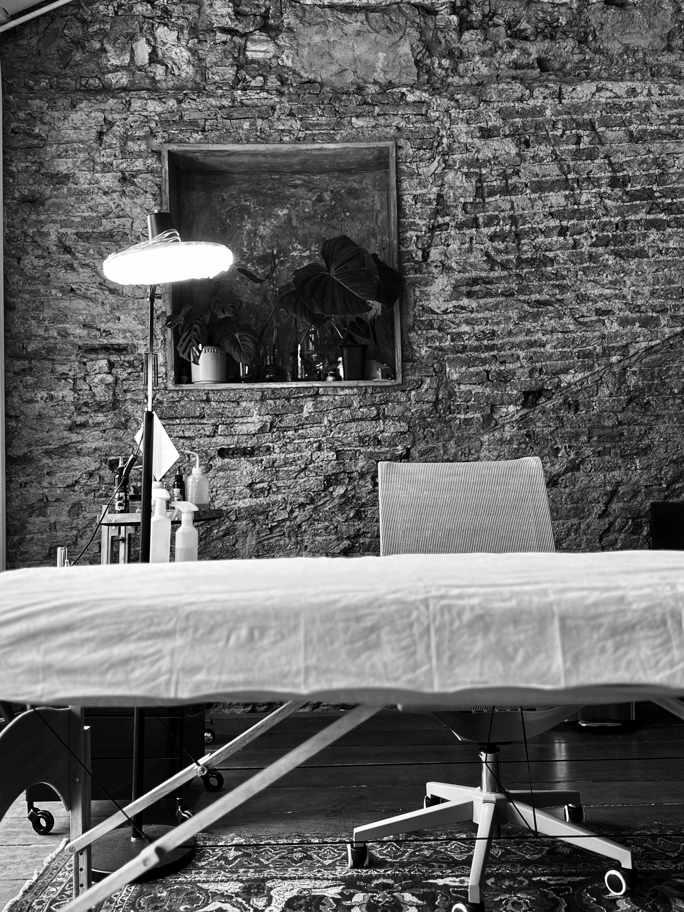 A room with an exposed brick wall, a mirror, several houseplants, a lamp, bottles, and a desk on a patterned rug, with a chair at the desk.