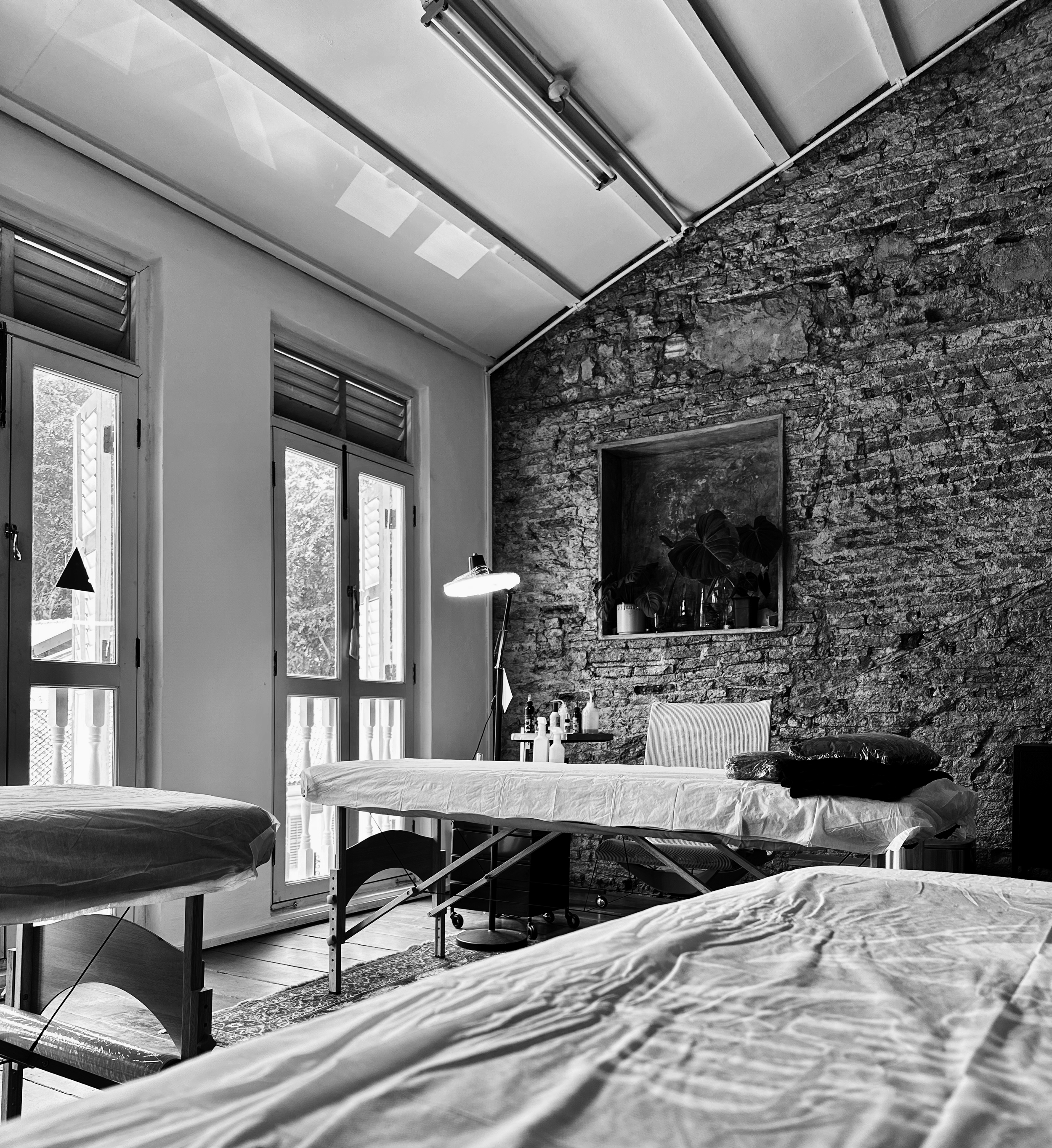 A black and white photo of a room with large windows, exposed brick wall, and various hygienic tattoo equipment including exam tables, a lamp, and supplies. The space appears to be an office or clinic setting.