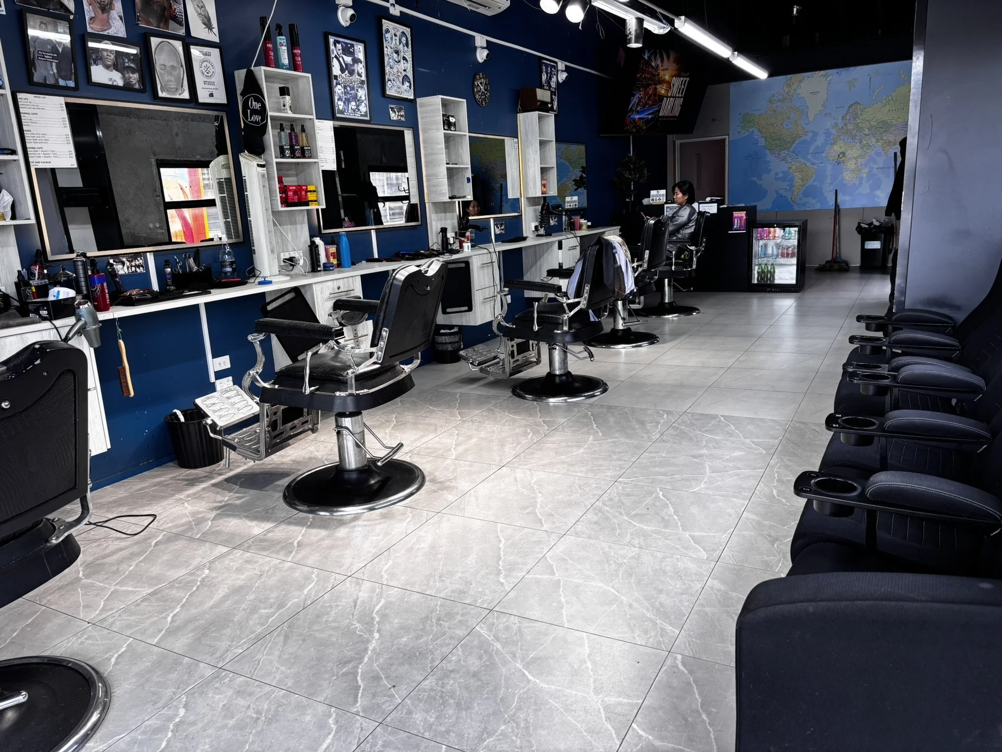 An empty barbershop with several styling chairs, mirrors on the wall, a world map poster, and a few customers and staff.