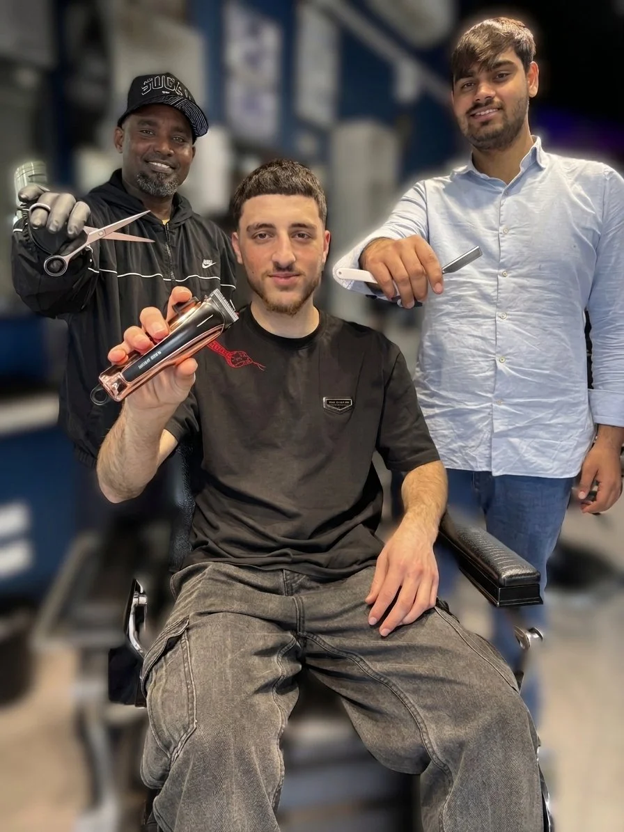 A man sitting in a barber chair holding a hair clipper, with two men standing behind him in a barbershop. The man on the left is holding scissors, and the man on the right is holding a straight razor.