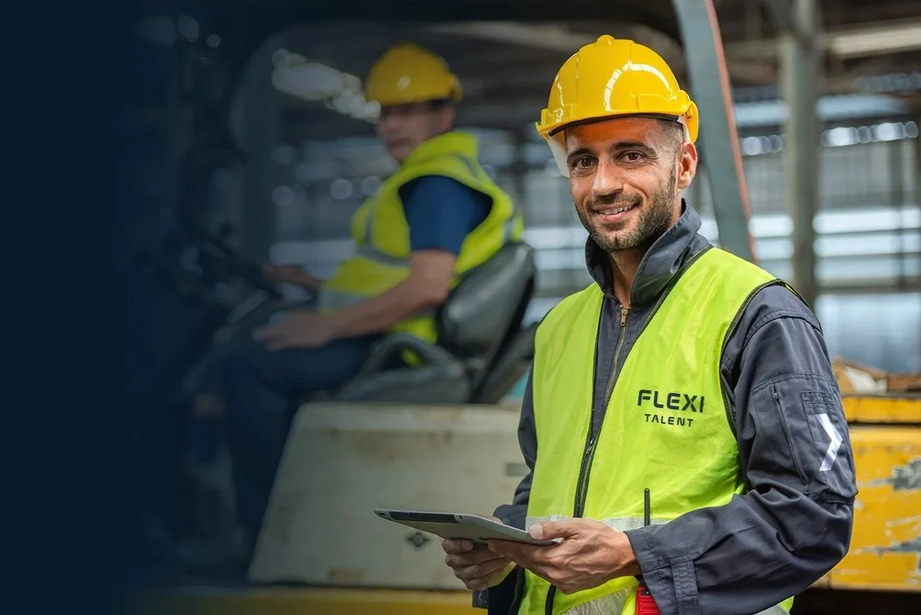 Two construction workers wearing yellow safety helmets and high-visibility vests on a construction site. One worker is smiling and holding a tablet, while the other is in a vehicle in the background.