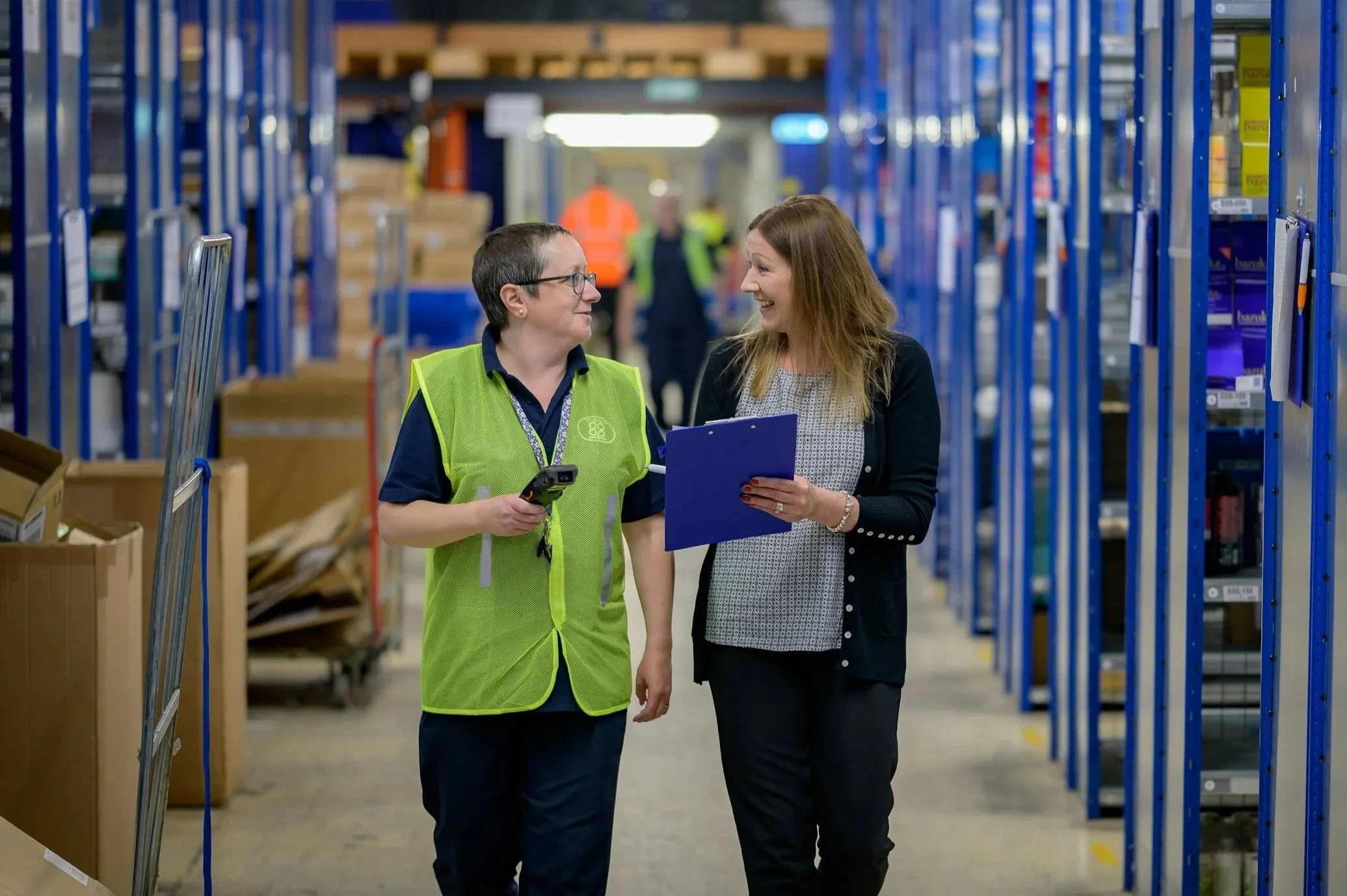 Two women are walking and talking in a warehouse or storage facility with metal shelves and boxes around. One woman, wearing glasses and a green safety vest, is holding a scanner. The other woman has long hair and is holding a clipboard. They are smiling at each other.
