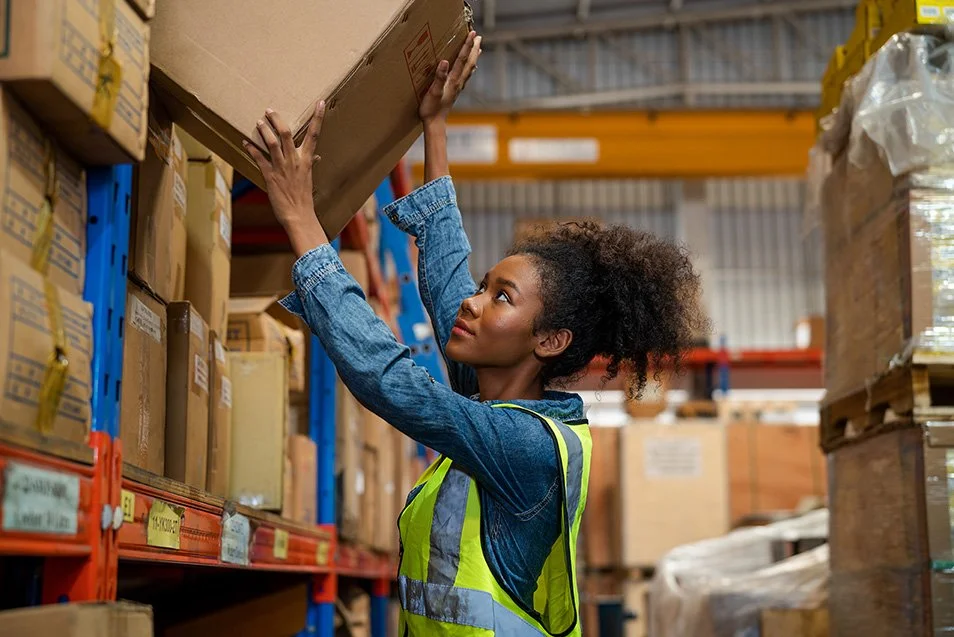 A woman in a high-visibility safety vest reaching up to pick a box from a warehouse shelf filled with boxes.