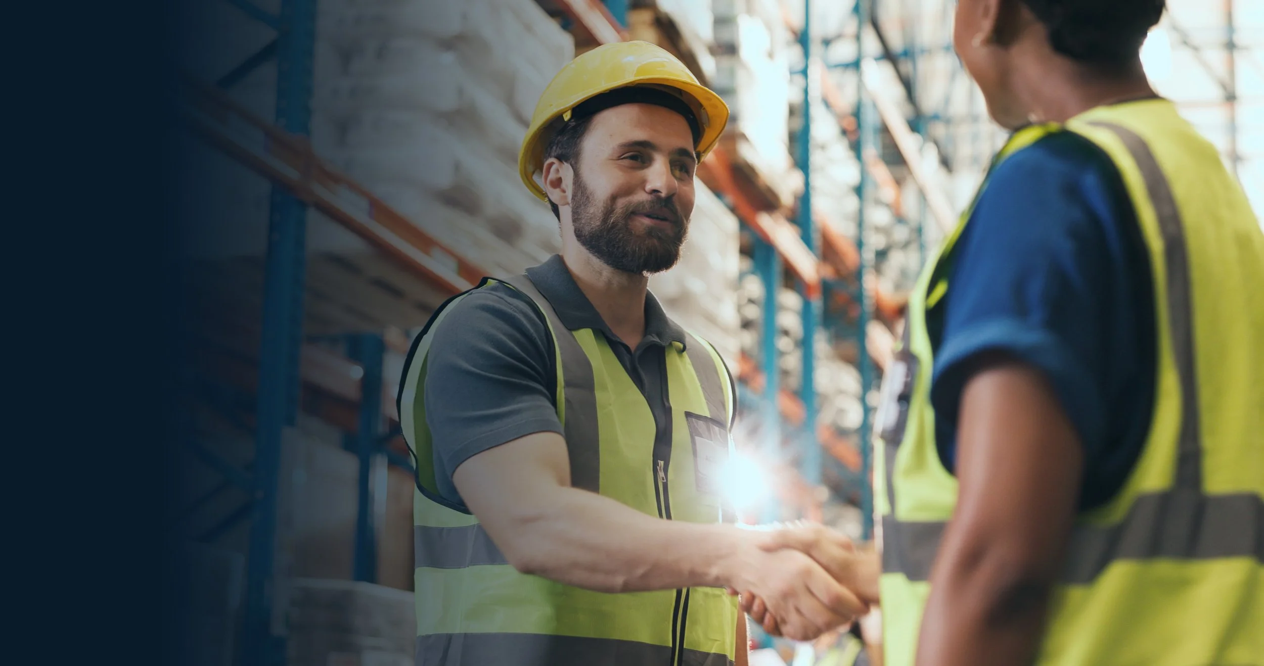 A man and a woman wearing safety helmets and vests shaking hands in a warehouse with shelves in the background.