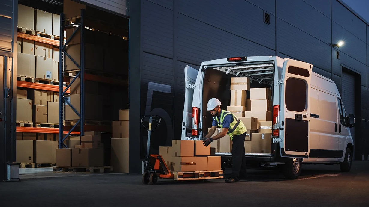 A worker in safety gear loading cardboard boxes onto a pallet in front of a delivery van at a warehouse.