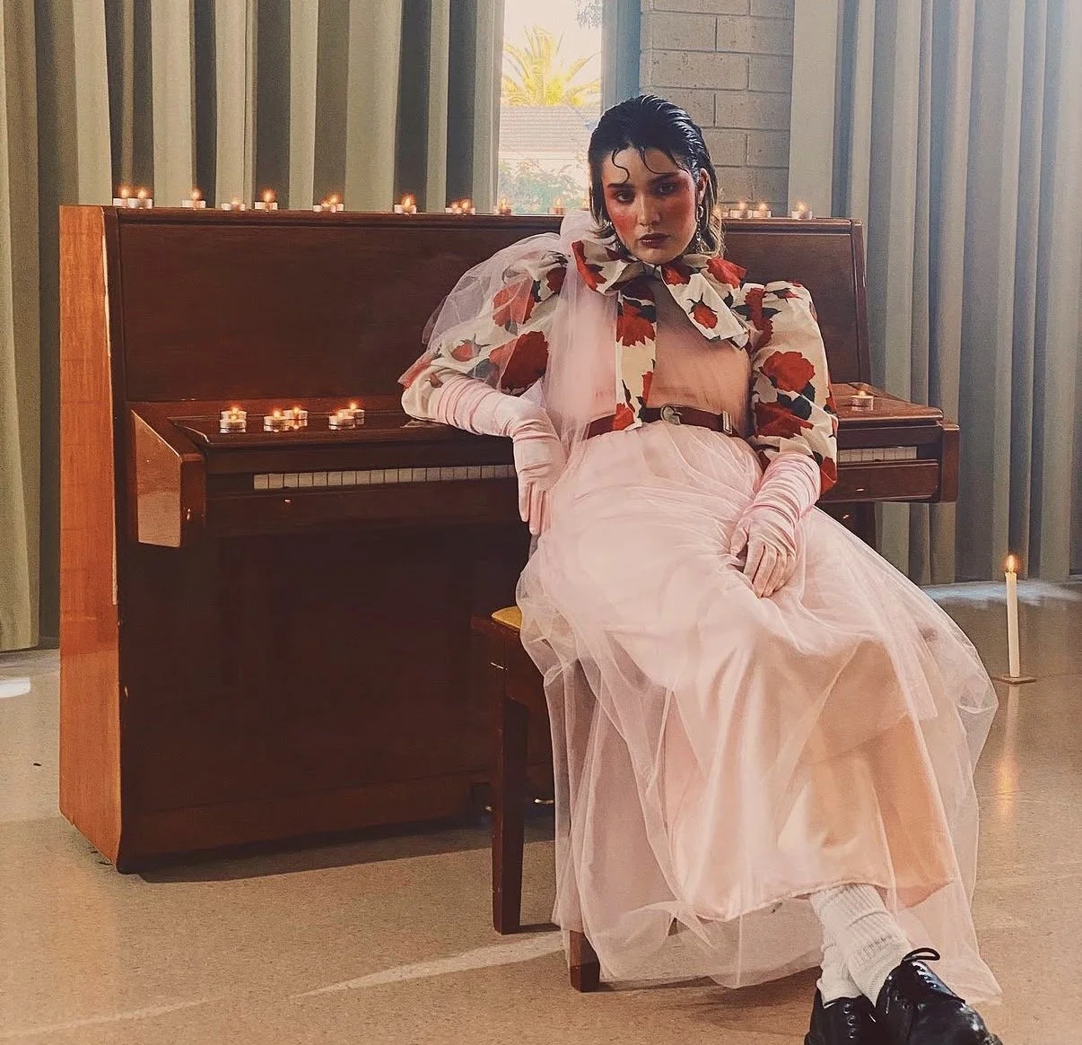A woman dressed in a vintage-inspired outfit with a floral jacket and pink tulle skirt, sitting next to a piano with lit candles in the background.