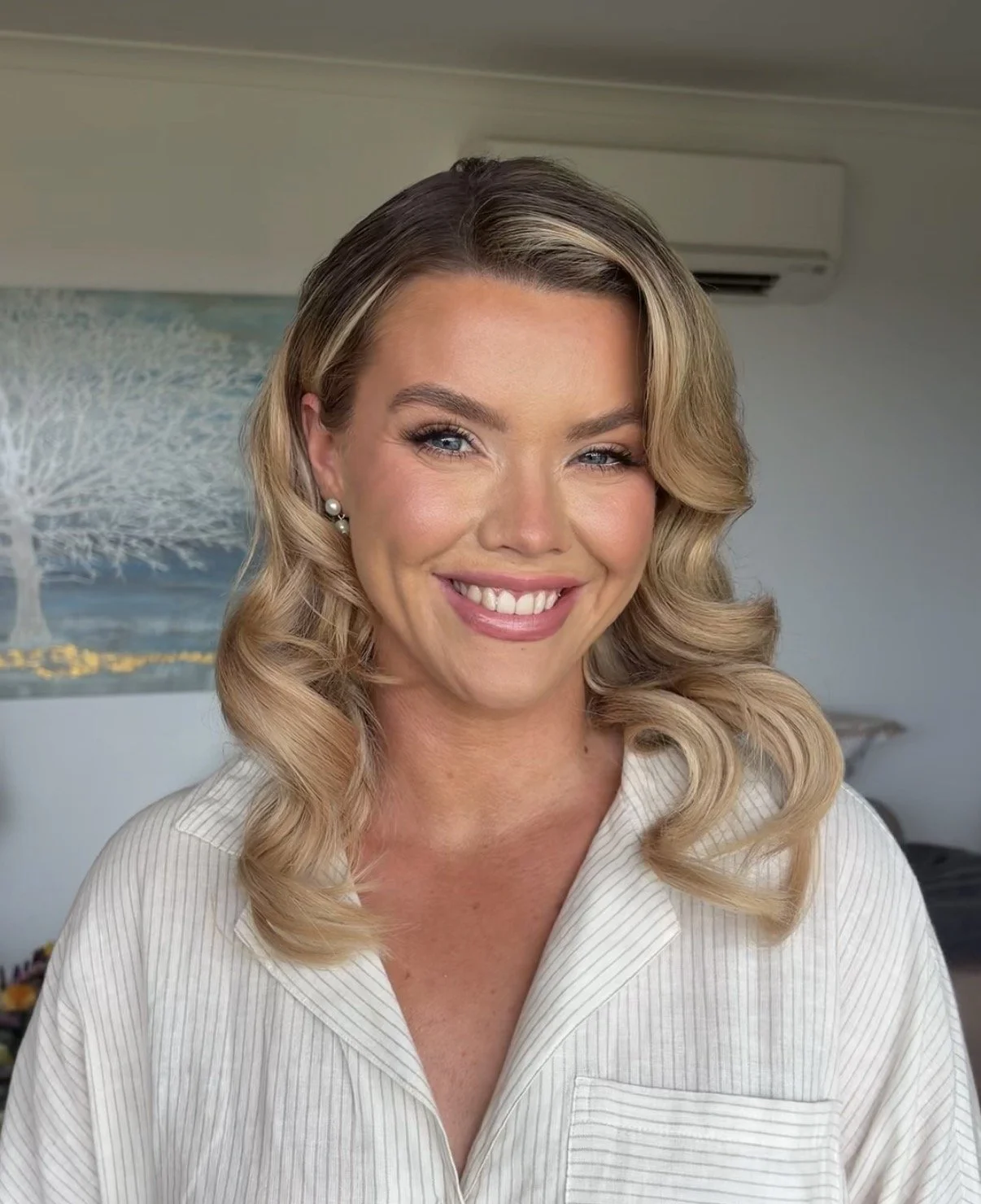 A woman with blonde, wavy hair, blue eyes, and a bright smile, wearing a white shirt and pearl earrings, standing indoors with a painting of a tree in the background.