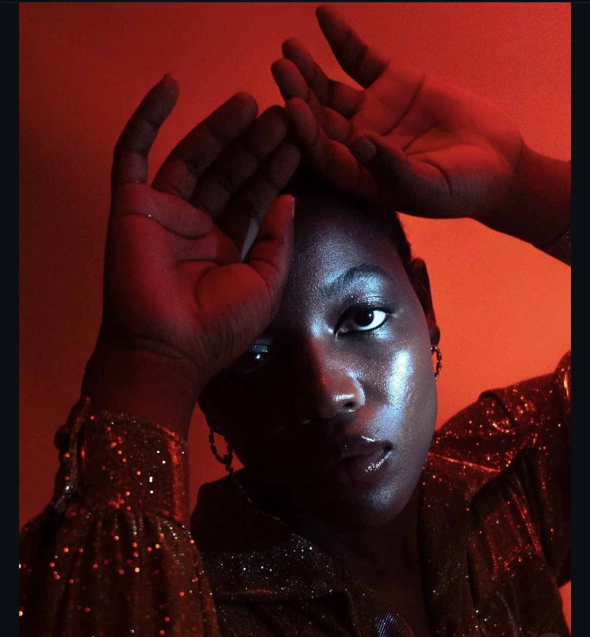 A young woman with dark skin and shiny makeup poses against a red background, with her hands raised above her head, framing her face. She is wearing earrings and a red sequined top.