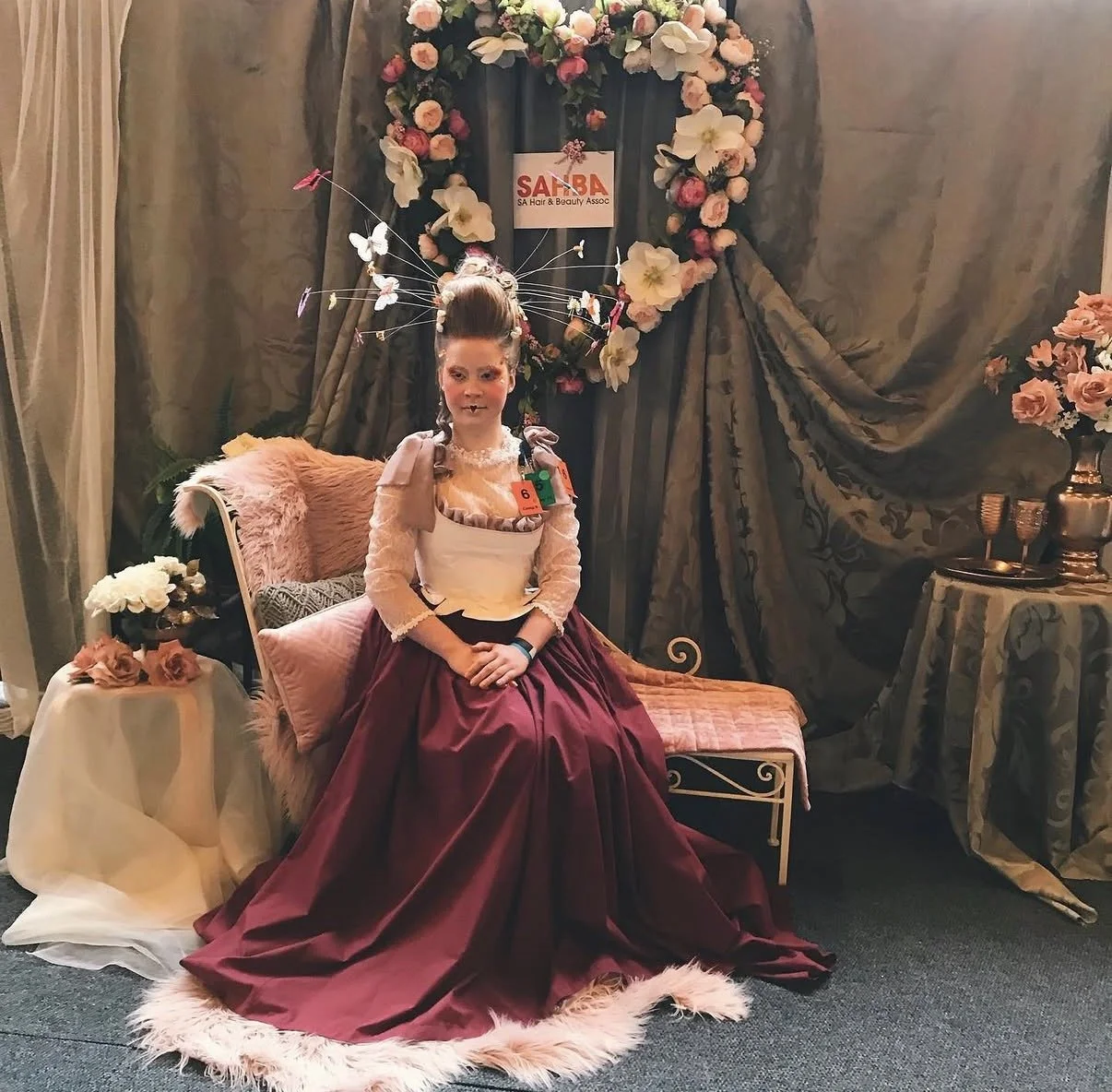 A young person sitting on a vintage-style bench in front of floral and fabric drapery, dressed in an elaborate costume with a beaded bodice, long burgundy skirt, and a halo-like headpiece with butterflies, during a themed event at SA Hair & Beauty Assoc.