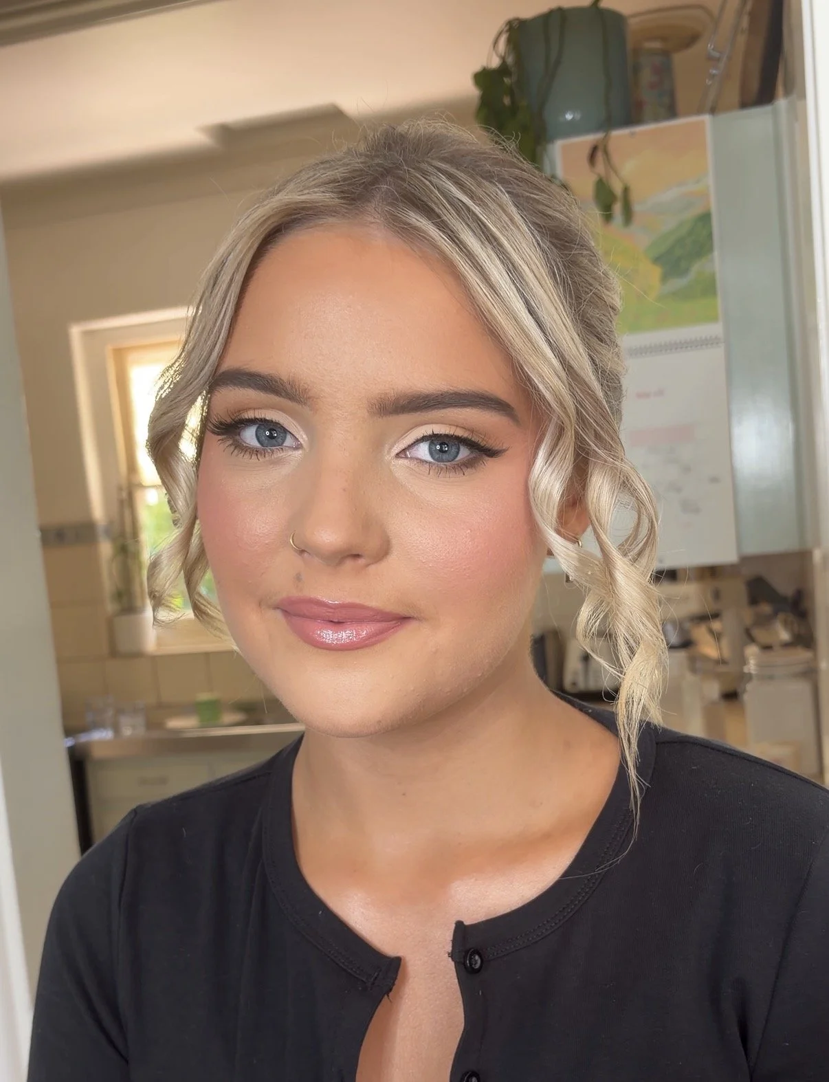 Close-up of a young woman with blonde, wavy hair, blue eyes, and makeup, wearing a black top in an indoor setting with a window, plant, and colorful painting in the background.