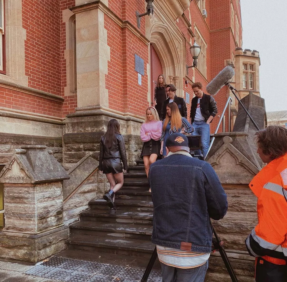 A film crew shooting a scene on the steps outside a historic red brick building with stone accents. Several young actors or extras are preparing to film, with one girl walking up the stairs and others standing at the top. A boom microphone is positioned above them, and two crew members are managing equipment and recording.