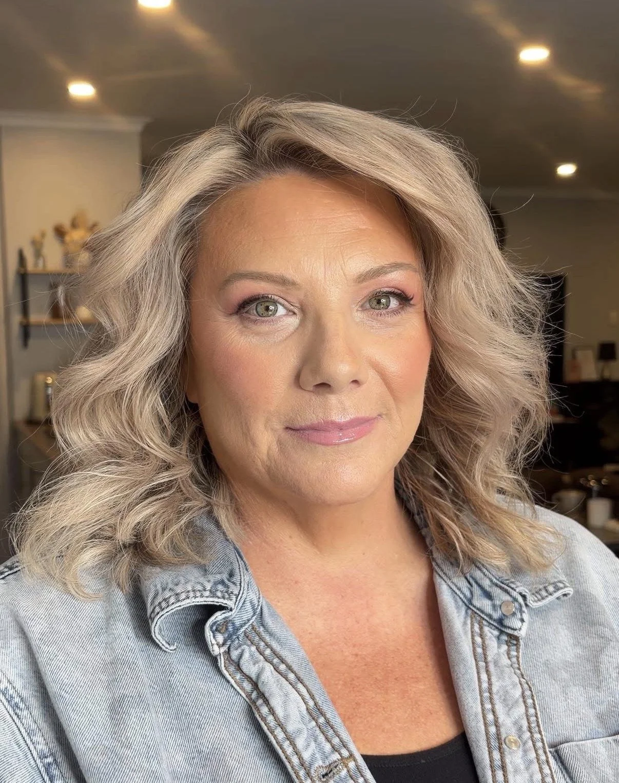 Close-up of a middle-aged woman with blonde, wavy hair and light makeup, wearing a denim jacket indoors.