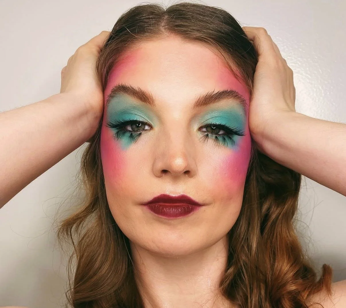 Close-up of a woman with colorful makeup, pink blush, teal eyeshadow, and dark lipstick, holding her head with both hands.