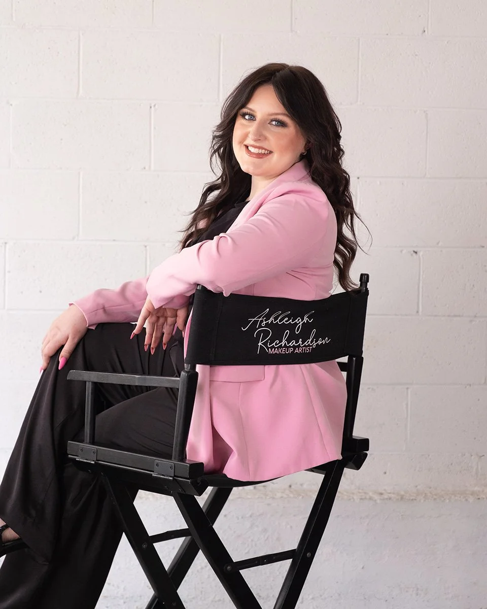 A woman with long dark hair and a pink blazer sitting on a black director's chair with embroidered white text that reads "Ashleigh Richardson Makeup Artist" against a white brick wall.