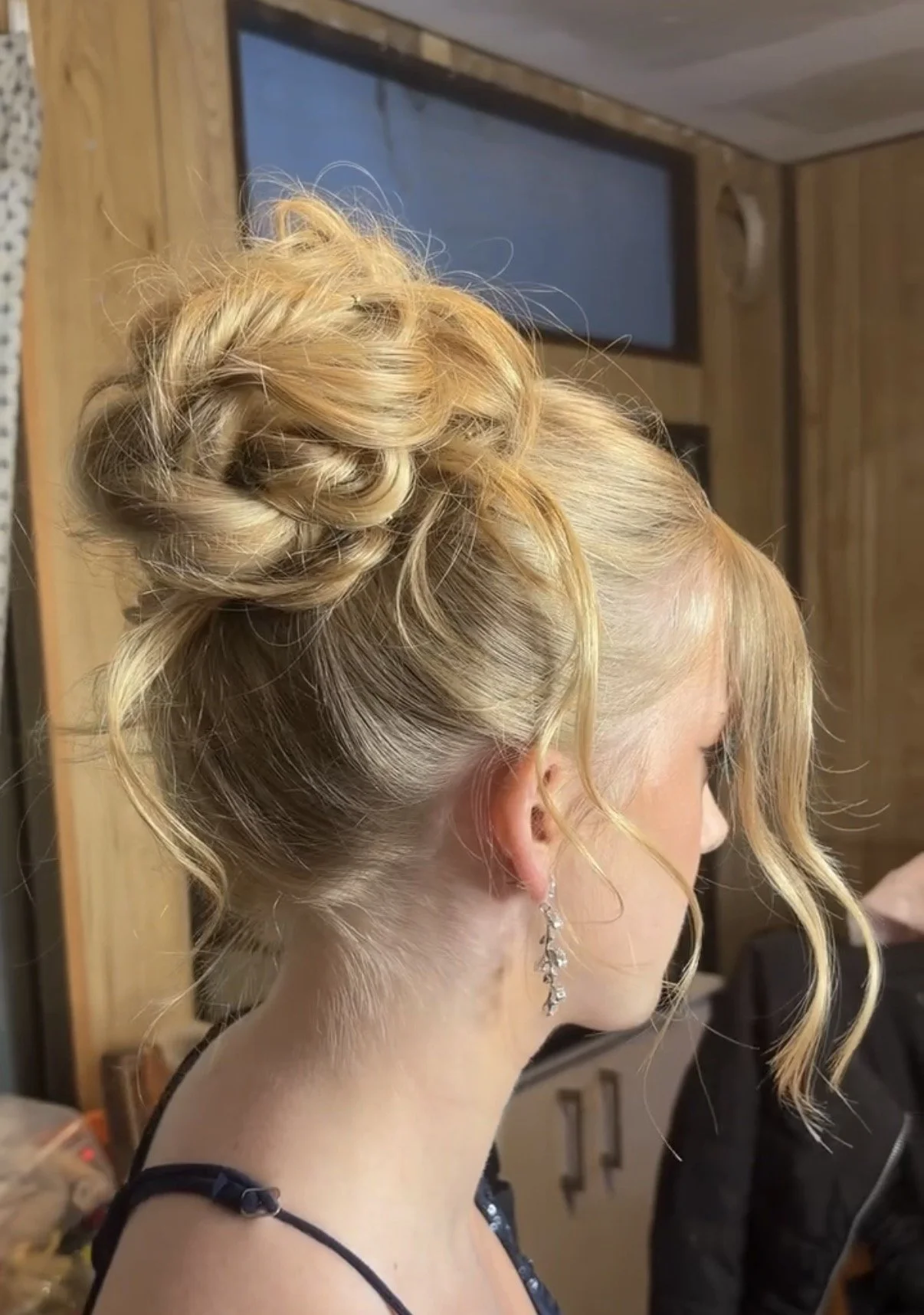 A woman with blonde hair styled in an intricate updo with curls and twists, wearing dangling earrings, and a black strap dress, in a room with wooden walls.