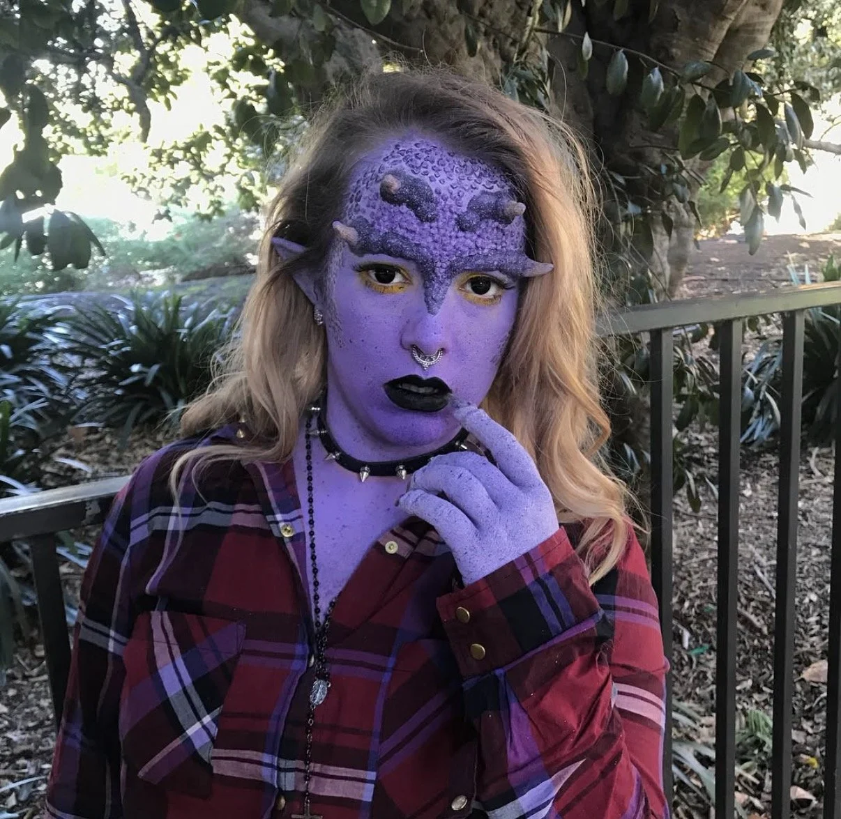 A person with purple alien-like facial makeup, wearing a maroon plaid shirt, earrings, a choker, and a necklace, posing outdoors near trees and a fence.