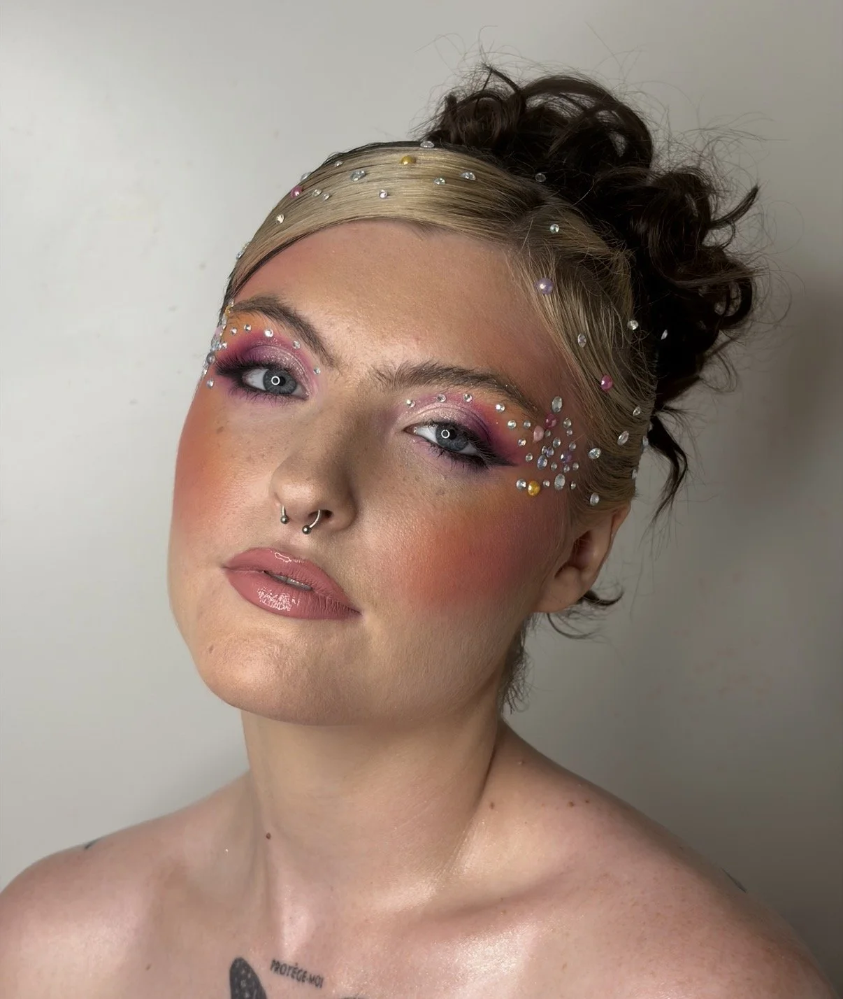 Portrait of a woman with dramatic makeup, decorated with various rhinestones around her eyes and on her head, with styled hair and a septum piercing.