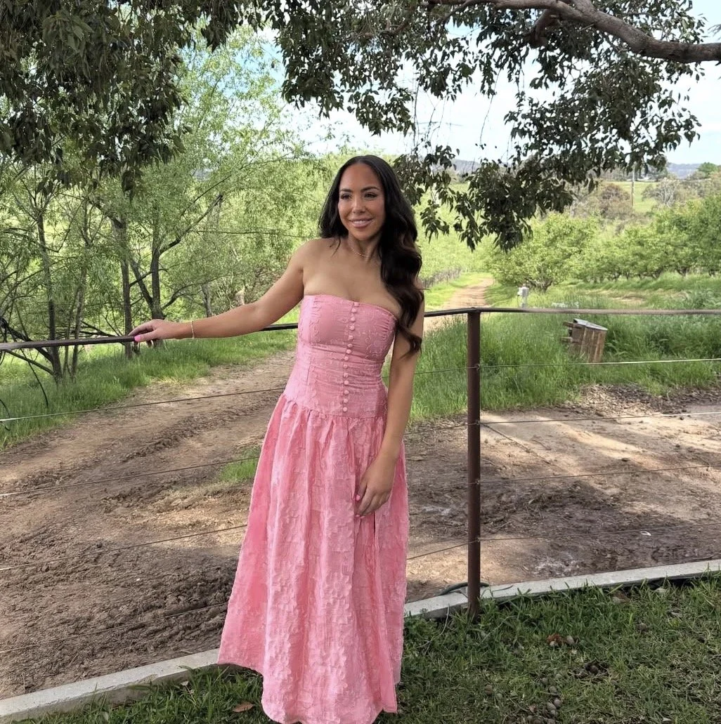 A woman in a strapless pink dress with a textured pattern, standing outdoors on a dirt pathway with trees and greenery in the background, holding a fence railing and smiling.