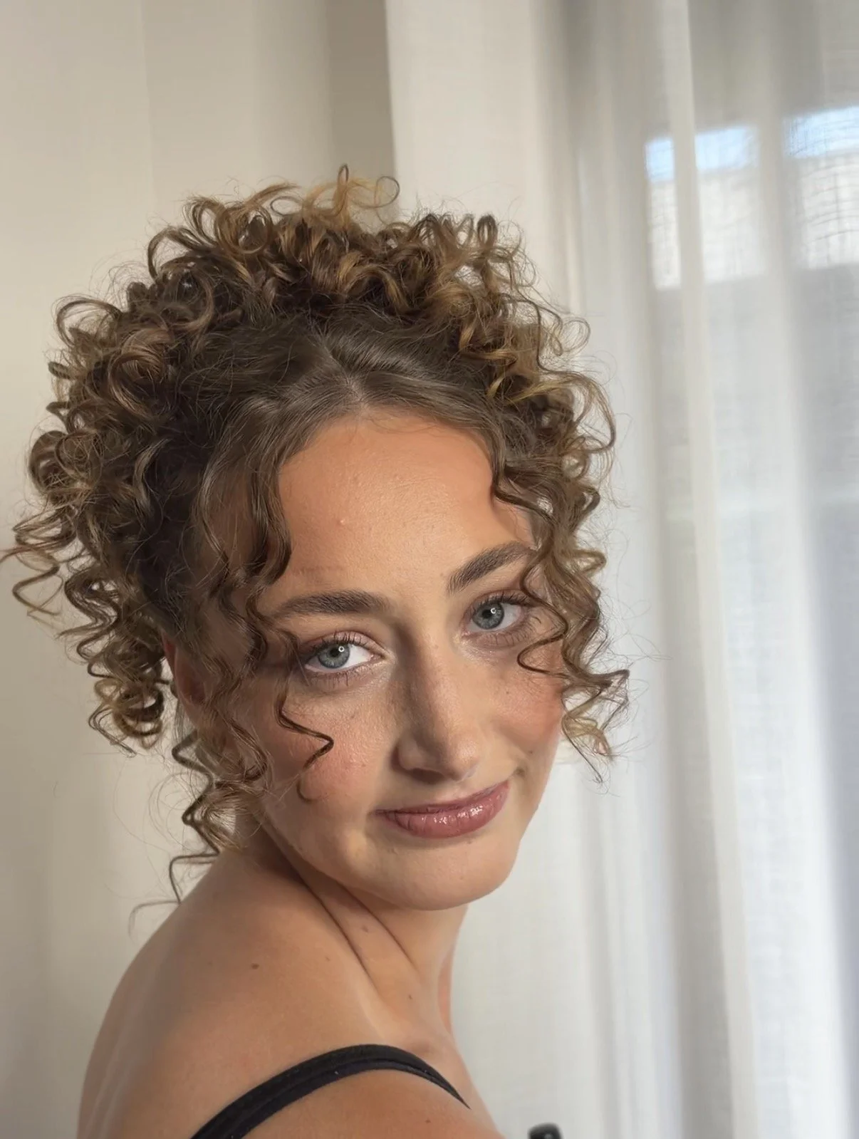 A woman with curly brunette hair and blue eyes. She is smiling and looking at the camera, with a sheer curtain and window in the background.