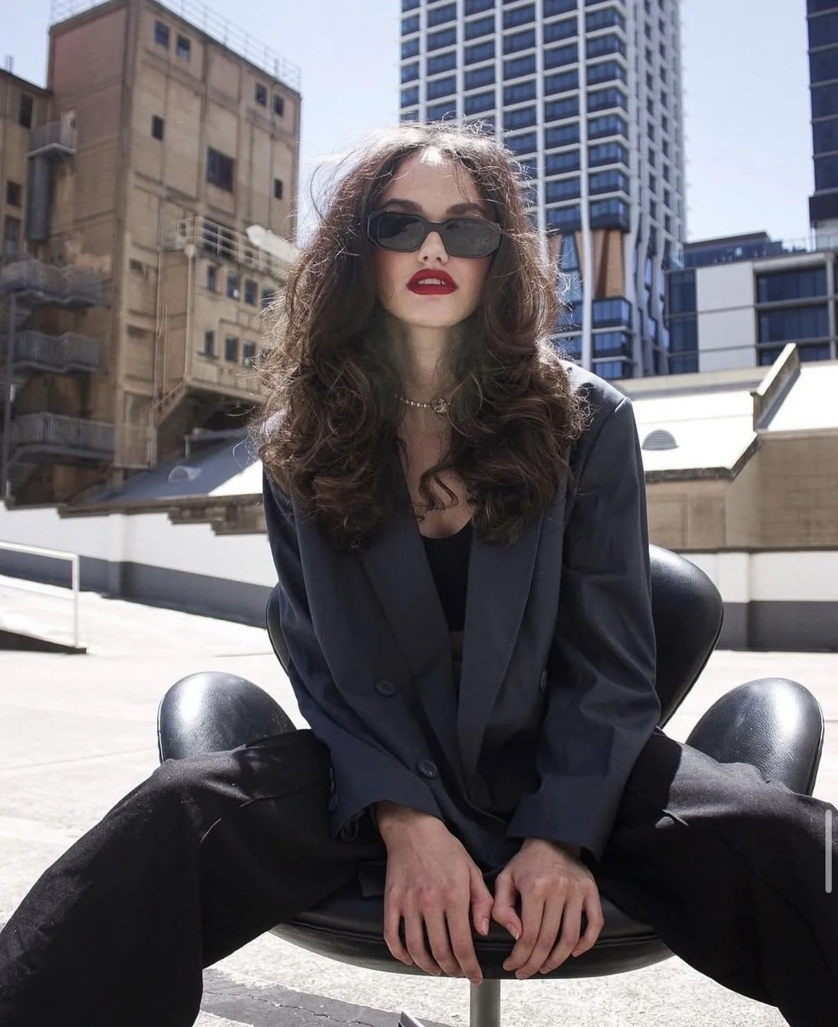 A woman with curly dark hair, wearing sunglasses, a black blazer, and red lipstick, sitting on a modern black chair outdoors with urban buildings and high-rise apartments in the background.