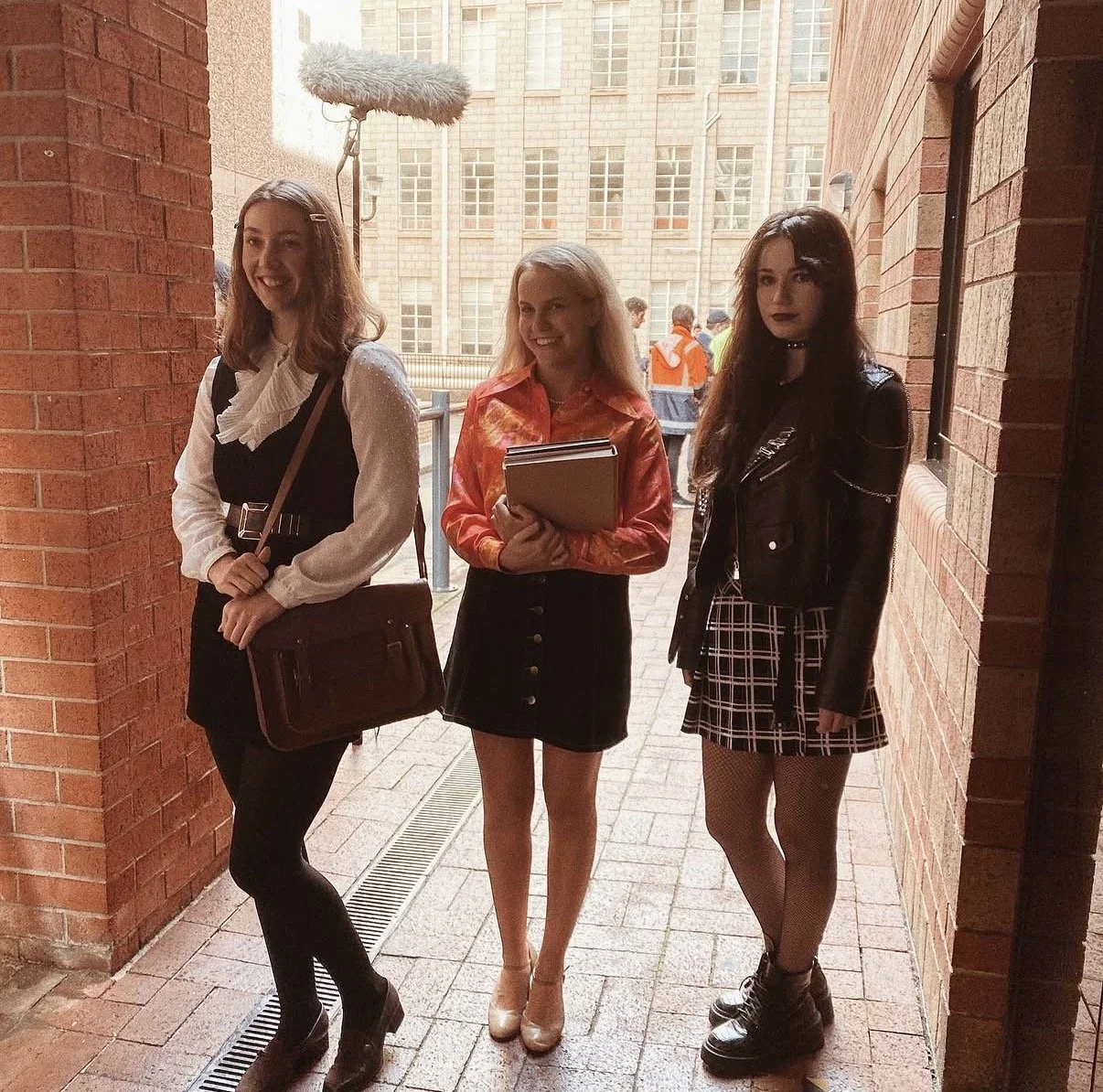Three young women standing in a brick corridor, smiling, with a city building in the background. The woman on the left is holding a bag and has a fluffy mic above her. The woman in the middle is holding notebooks and wearing a colorful jacket. The woman on the right is dressed in a black leather jacket, plaid skirt, and fishnet stockings.