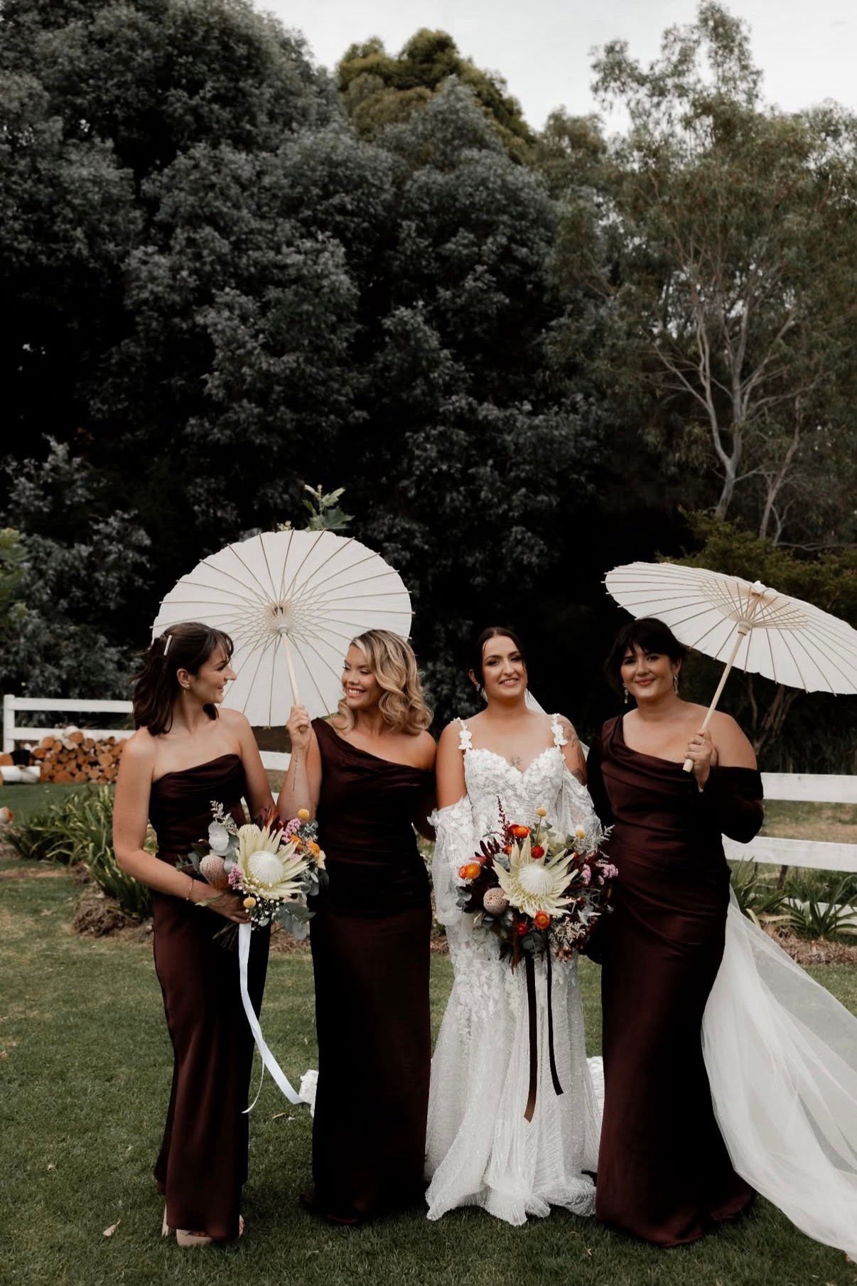 Four women in formal attire, one in a white wedding dress and three in matching dark brown dresses, stand outdoors on grass with trees in the background. Two women hold white parasols, and all hold bouquets of flowers, smiling and posing together.