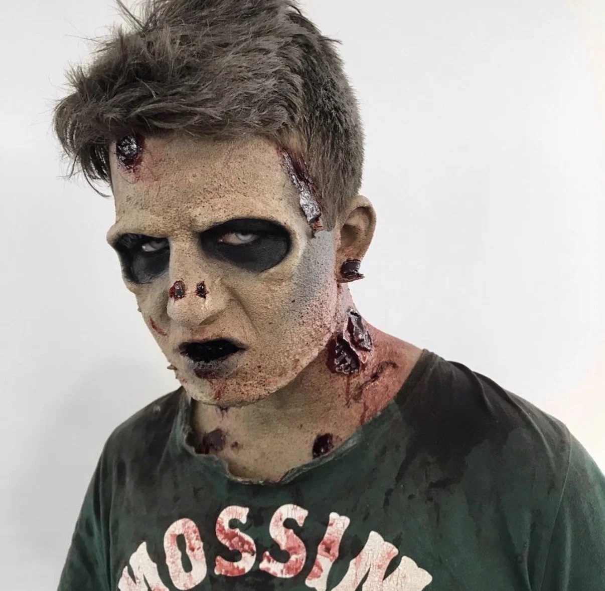 Person with zombie makeup, black eye circles, fake wounds, and wounds with blood; wearing a dark green T-shirt with white and red lettering