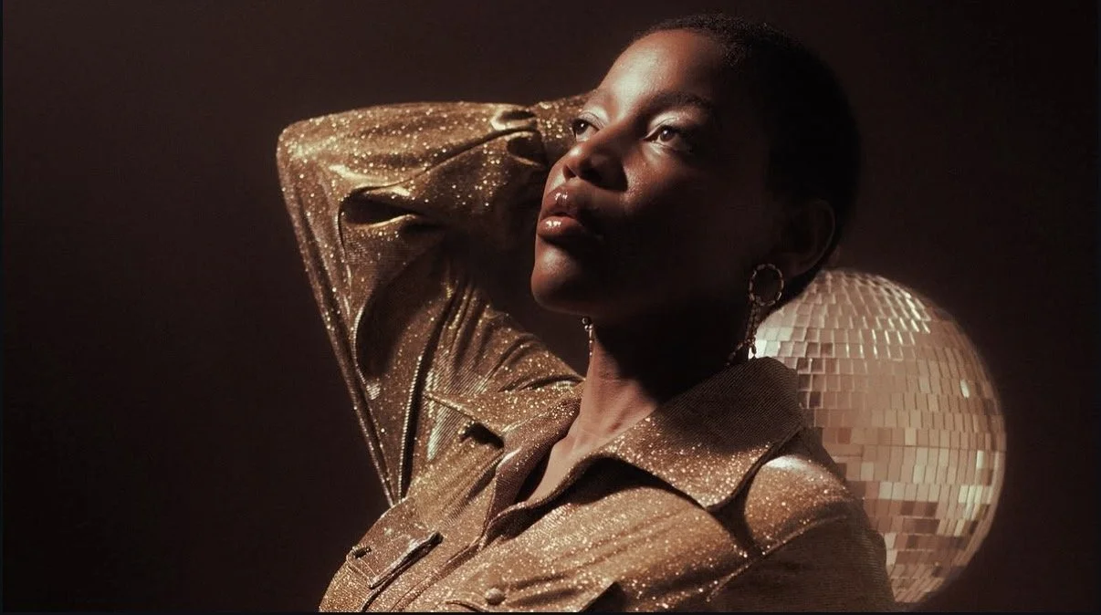 A woman with short hair and hoop earrings posing with her hand behind her head, wearing a sparkly gold jacket, with a disco ball in the background.