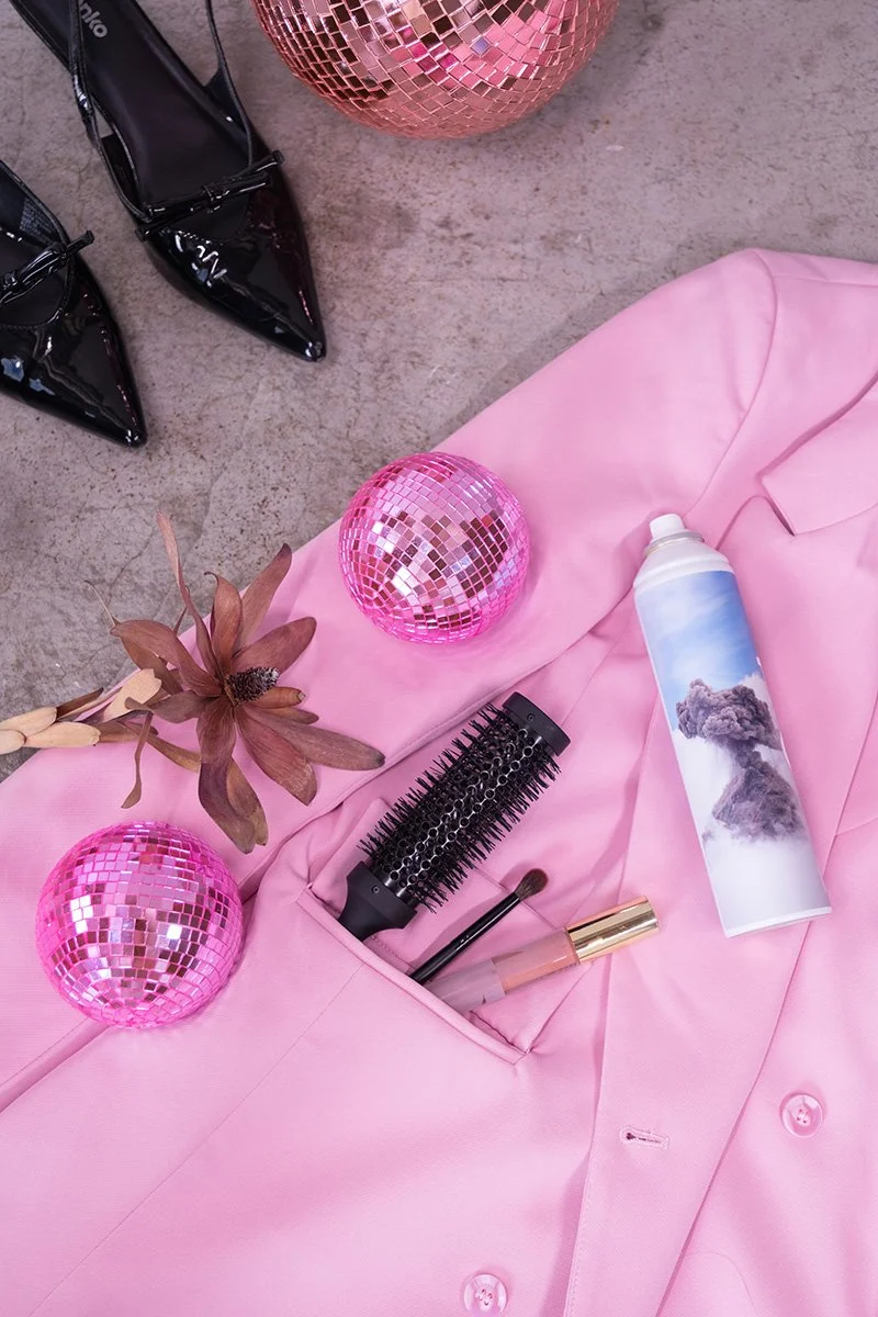 A pink blazer, black patent high heels, pink disco balls, a dried flower, a hairbrush, a makeup brush, and a water spray bottle with a mountain scene, all arranged on a concrete floor.