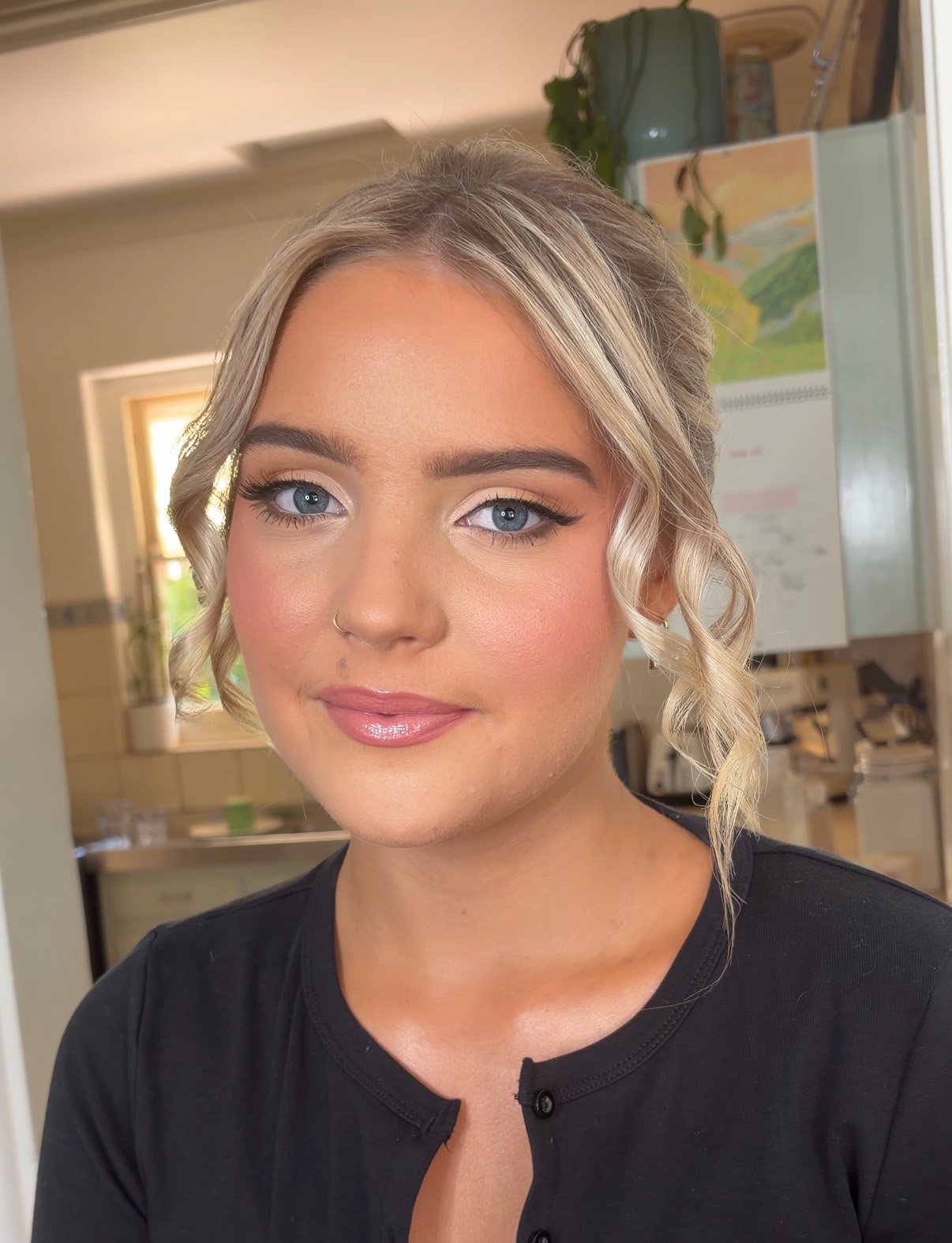 Close-up of a young woman with blonde hair styled in loose curls, wearing makeup, a nose ring, and a black top, in a kitchen with a window, plants, and a calendar in the background.
