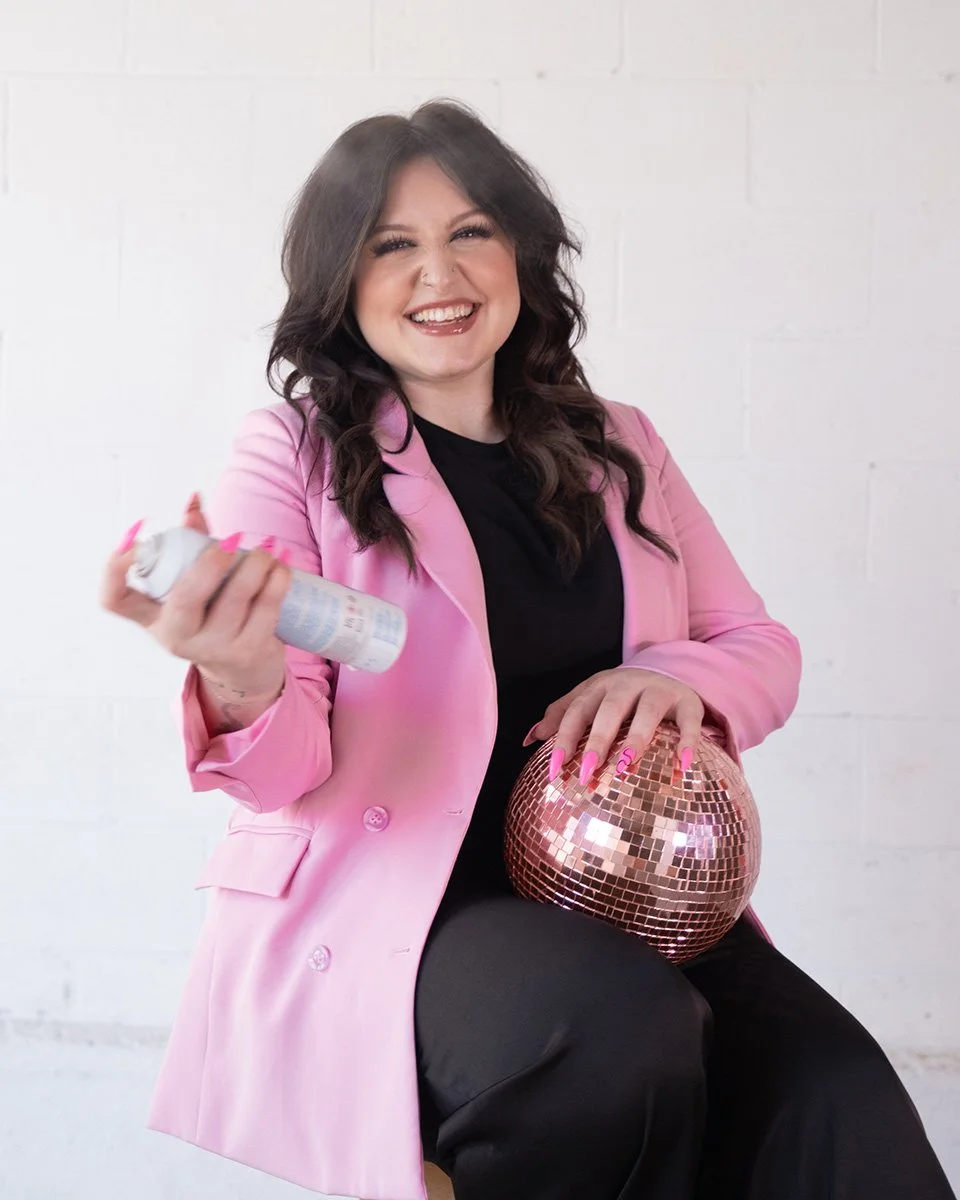 A woman with dark, wavy hair, wearing a pink blazer and black top, smiling and holding a spray can in one hand and a rose gold disco ball on her lap, sitting against a plain white wall.
