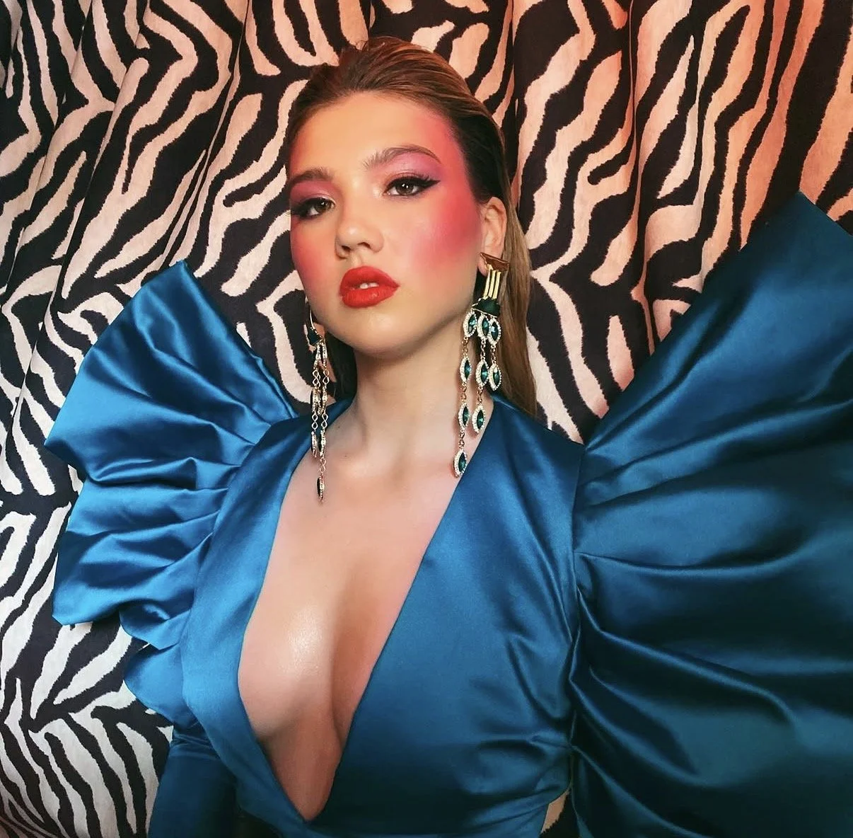A woman wearing a vibrant blue satin dress with dramatic ruffled sleeves, posing against a zebra-print backdrop. She has makeup with bold red lipstick, pink blush, and eye makeup, and is accessorized with large, ornate earrings featuring green and gold details.