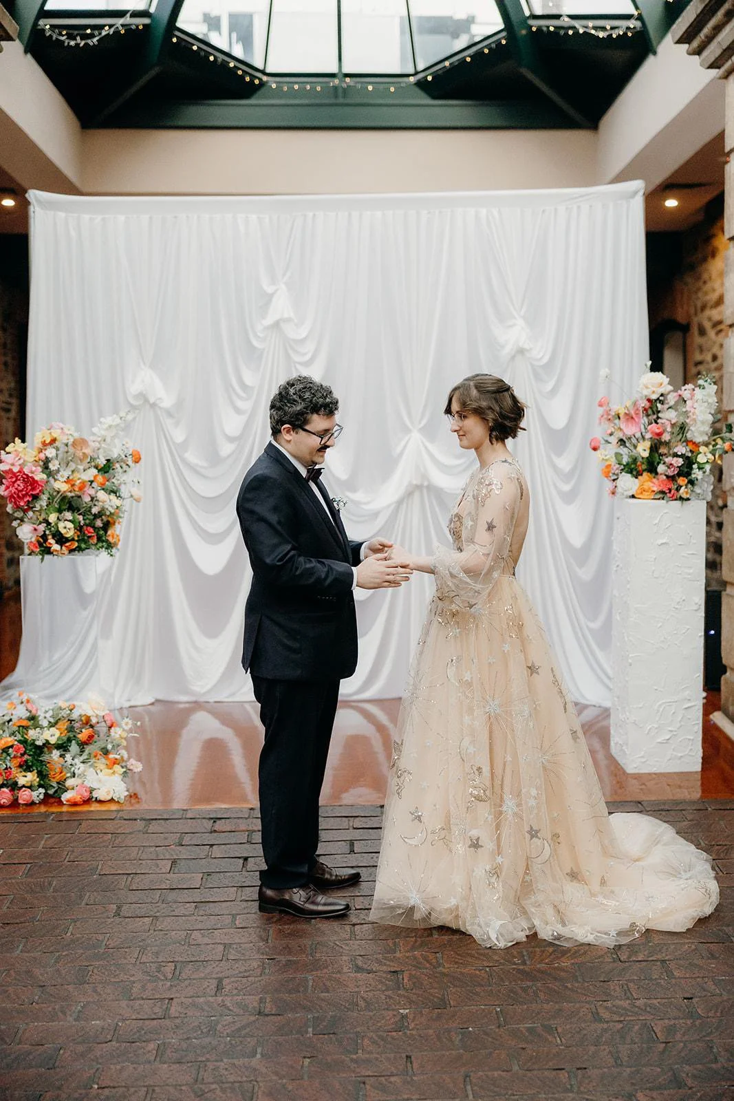 A wedding ceremony with a couple exchanging rings in front of a white draped backdrop with floral arrangements on either side.